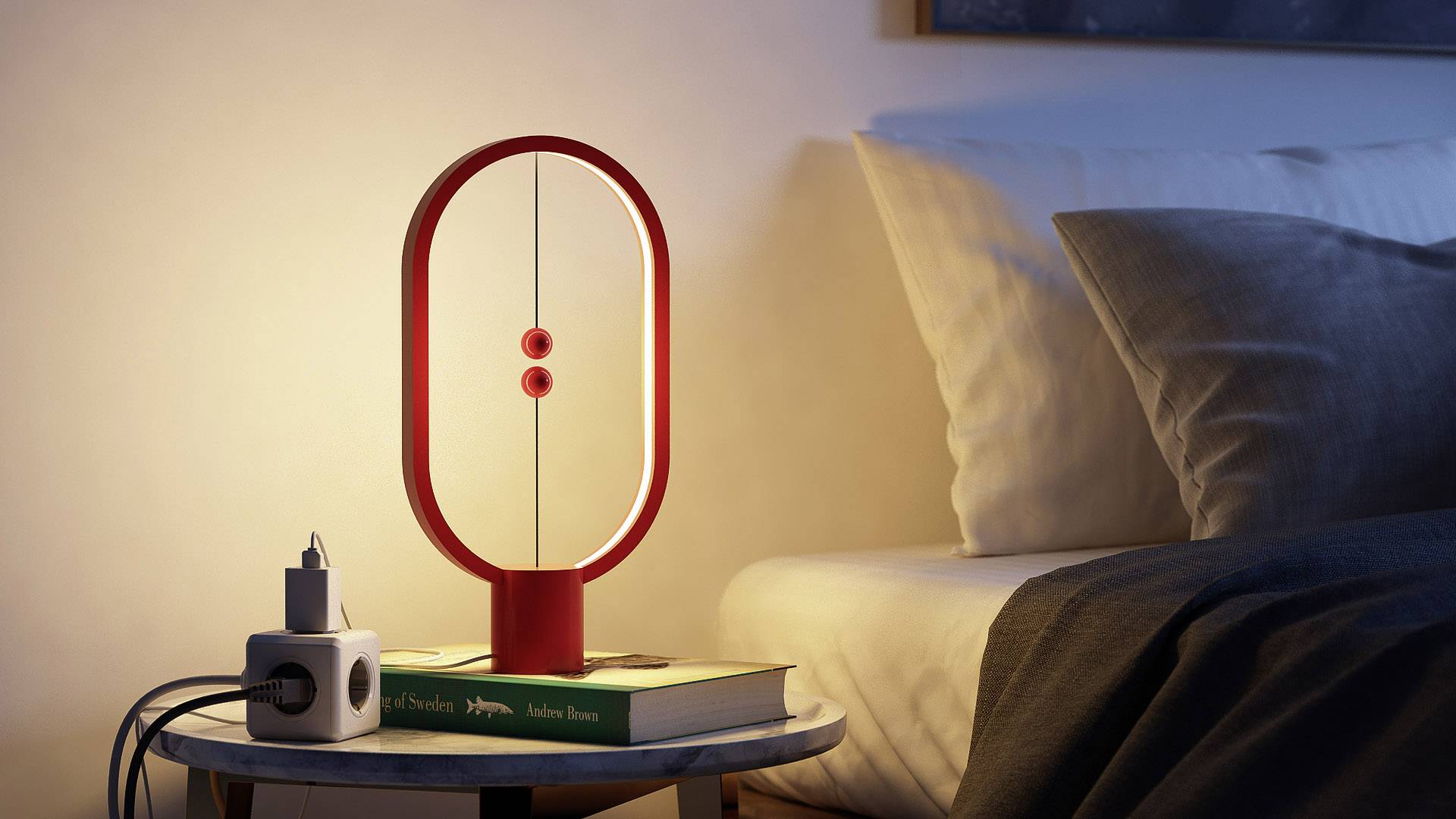 Eine rote Tischlampe steht eingeschaltet auf einem runden Tisch neben einem Bett. Darunter liegt ein Buch. Ein Ladegerät ist ebenfalls zu sehen.