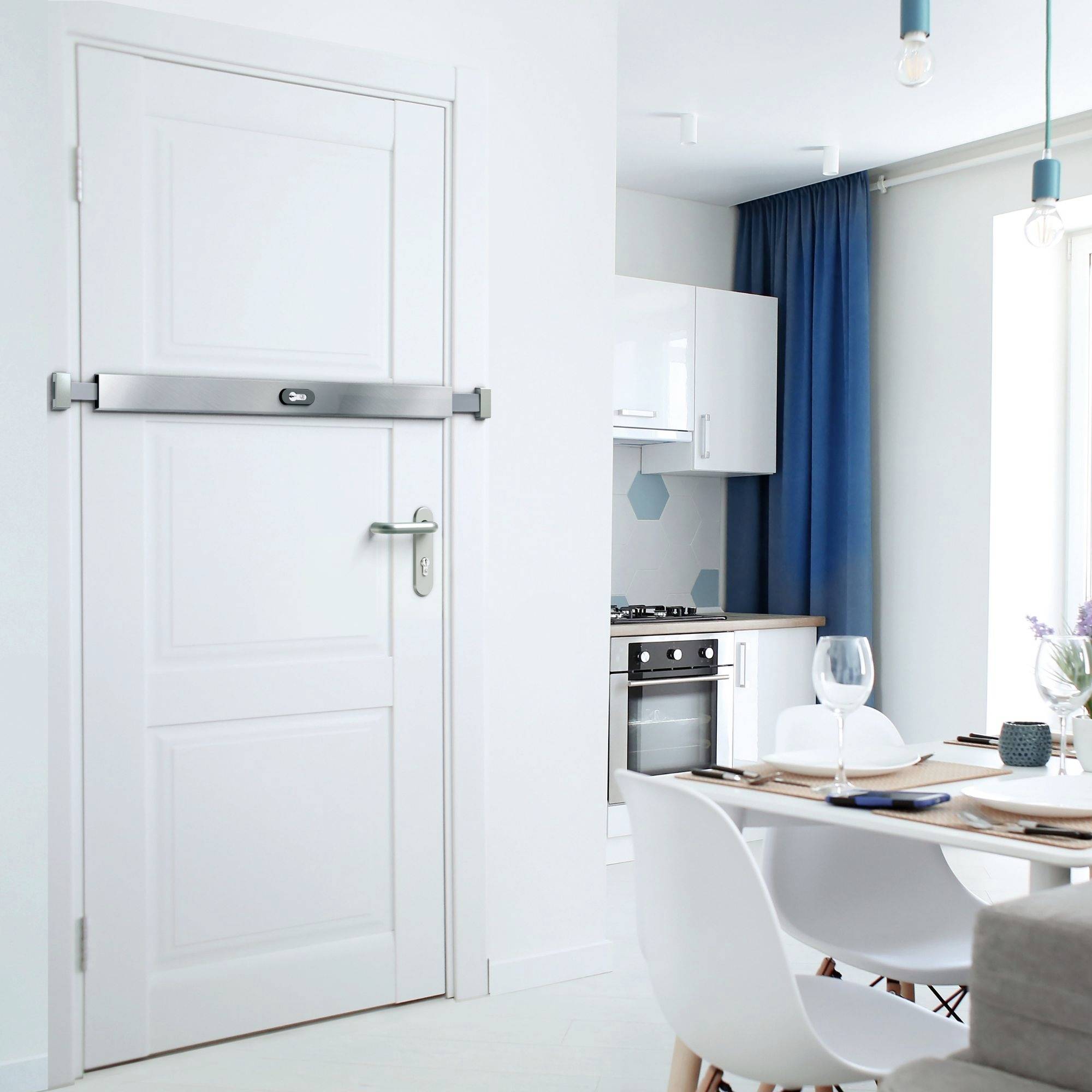 Eine weiße Wohnungstür mit einem silbernen Panzerriegel ist im Vordergrund prominent. Sie scheint in einen Küchen-/Essbereich zu führen. Der Küchen-/Essbereich ist hinter der Tür sichtbar und verfügt über weiße Schränke, einen für eine Mahlzeit gedeckten Esstisch und einen blauen Vorhang.