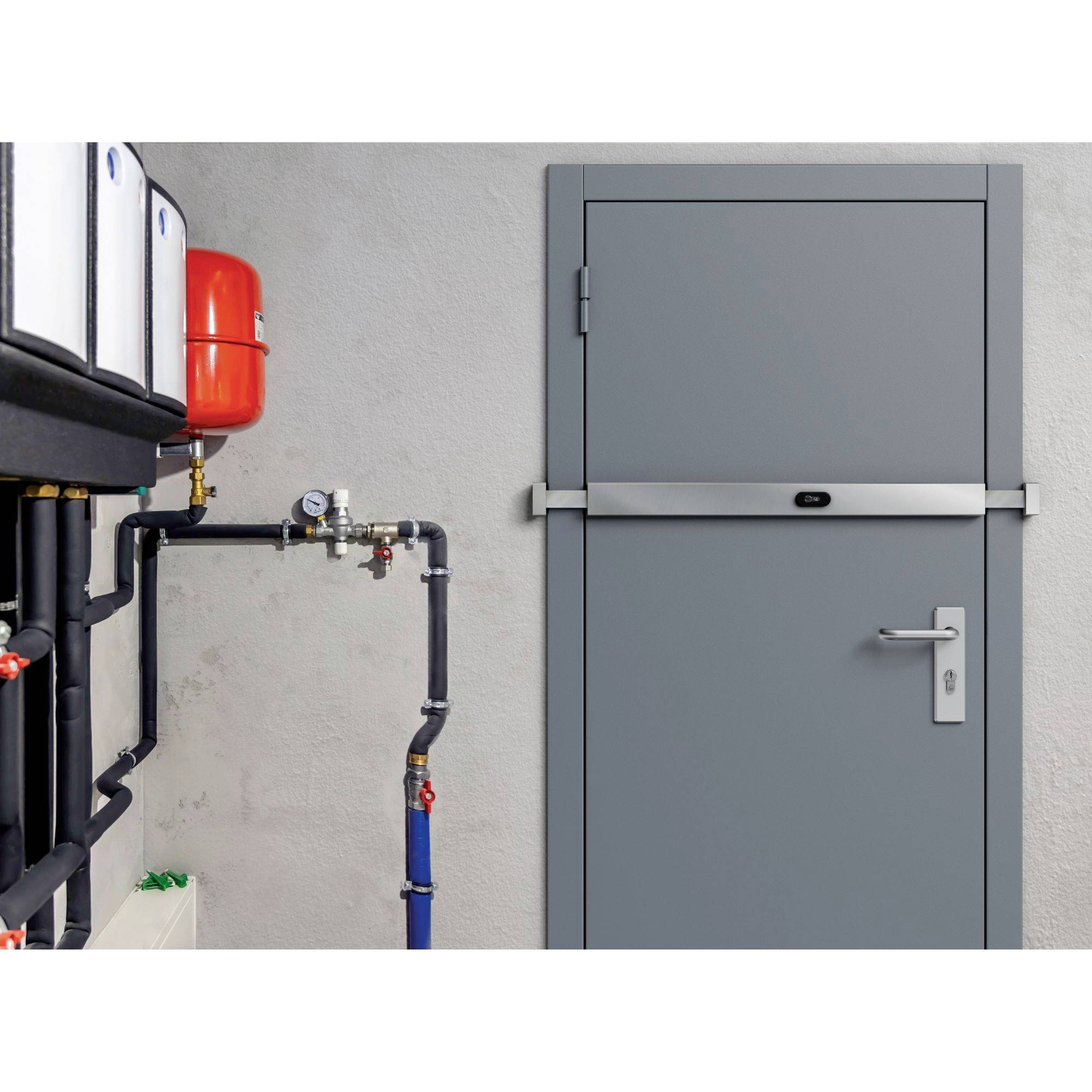 Das Bild zeigt eine graue, massive, industriell wirkende Tür und einen Teil von Sanitärinstallationen vor einer hellgrauen Wand. Die Tür ist mittig mit einem horizontalen Panzerriegel aus Metall verschlossen. Die Sanitärinstallationen umfassen Rohre, einen Druckbehälter und ein Manometer.