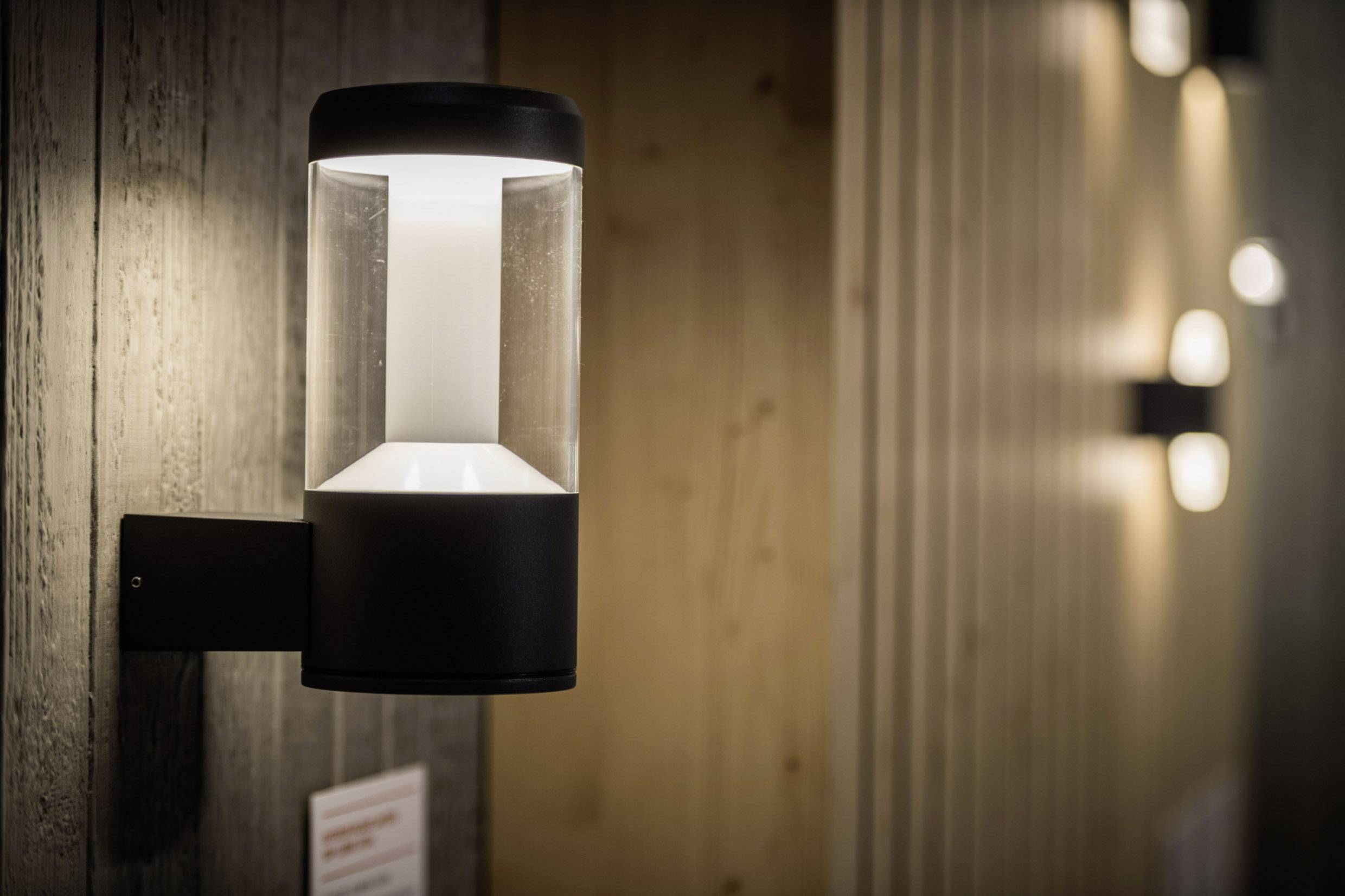 Moderne Wandlampe mit rundem, schwarzem Gehäuse, das diffuses Licht nach unten abstrahlt, an einer Holzwand montiert.