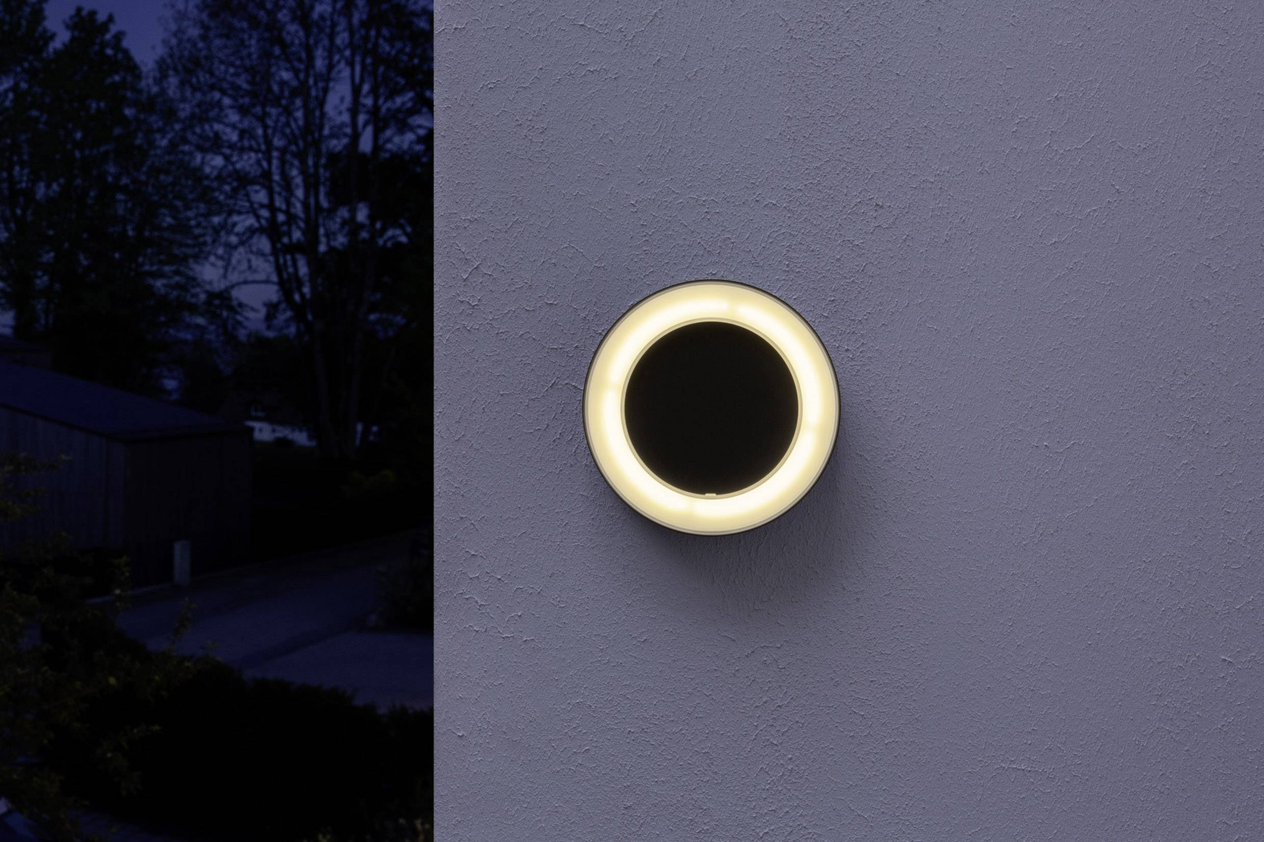Runde, leuchtende Außenlampe an einer Wand, beleuchtet einen Bereich in der Dunkelheit. Umgebung zeigt Bäume und ein Haus im Hintergrund.
