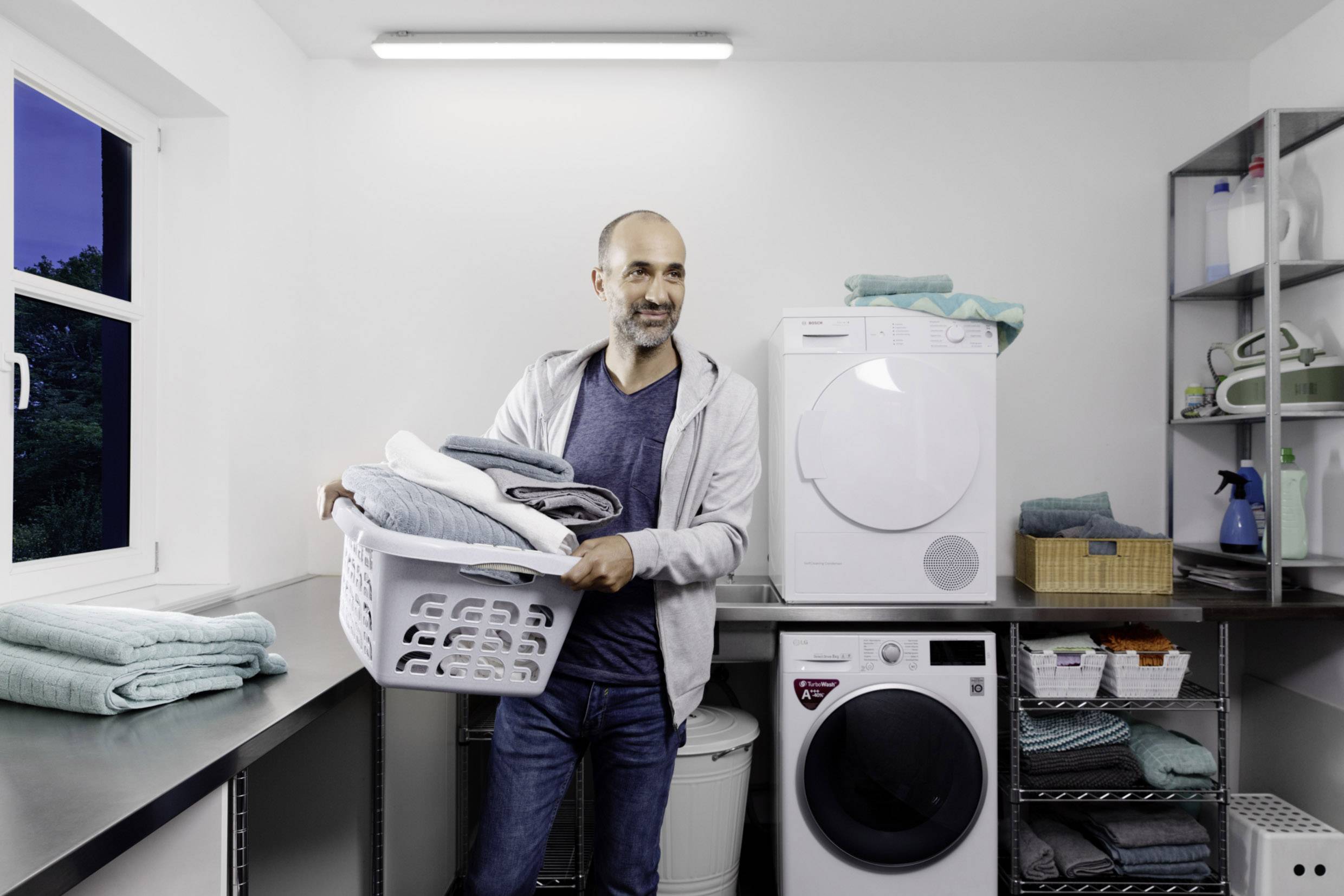 Ein Mann steht in einem Hauswirtschaftsraum und hält einen Wäschekorb mit gefalteten Handtüchern. Im Hintergrund sind Waschmaschine und Trockner zu sehen.