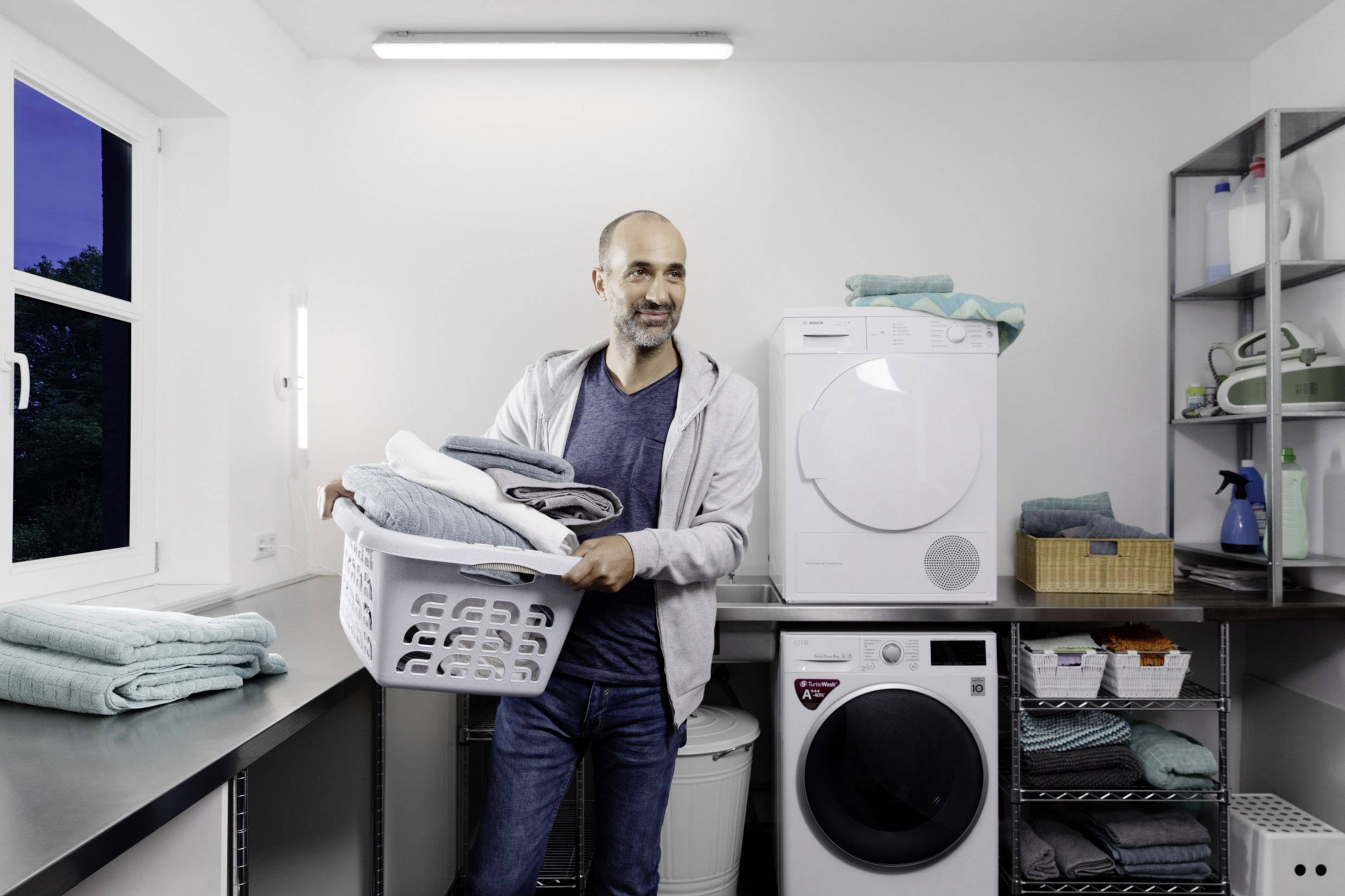 Eine Person hält einen Korb voller Wäsche in einem hellen Waschraum mit einem Wäschetrockner und einer Waschmaschine im Hintergrund.