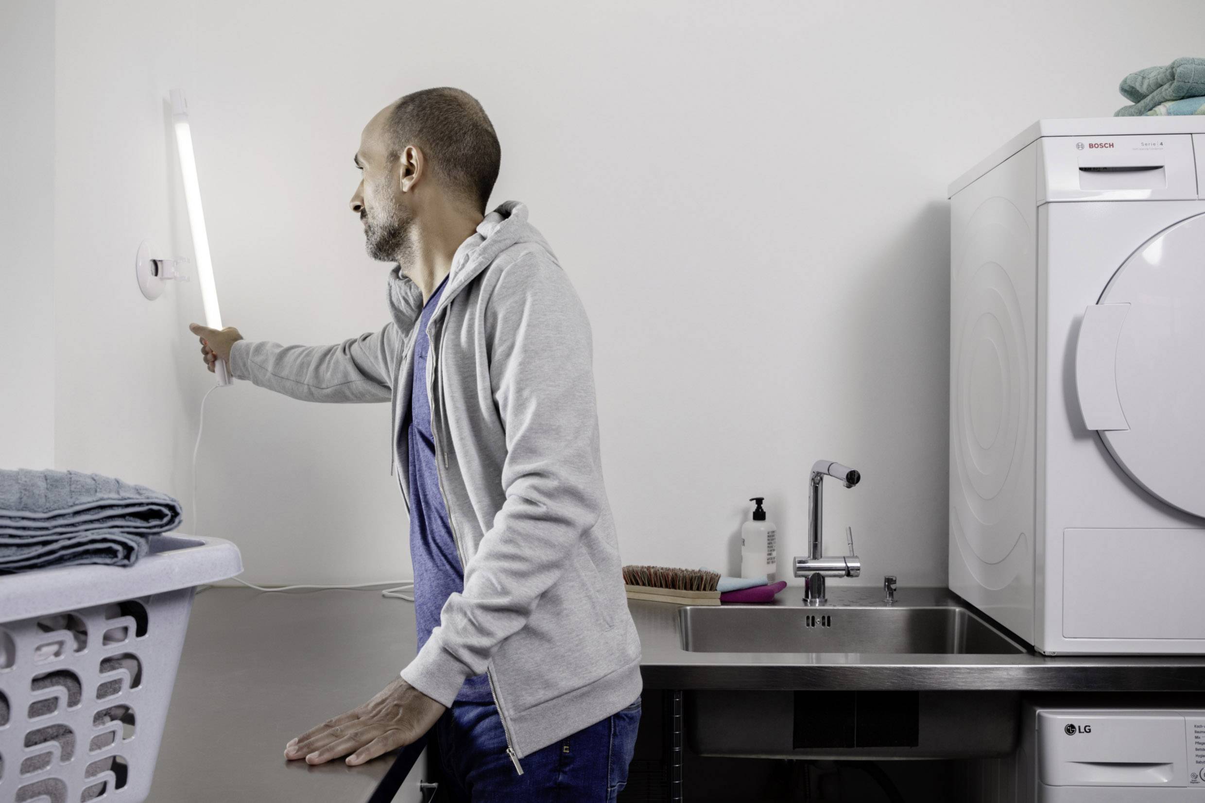 Ein Mann schaltet in einer Waschküche das Licht ein. Neben ihm befinden sich eine Waschmaschine, ein Trockner und ein Waschbecken.