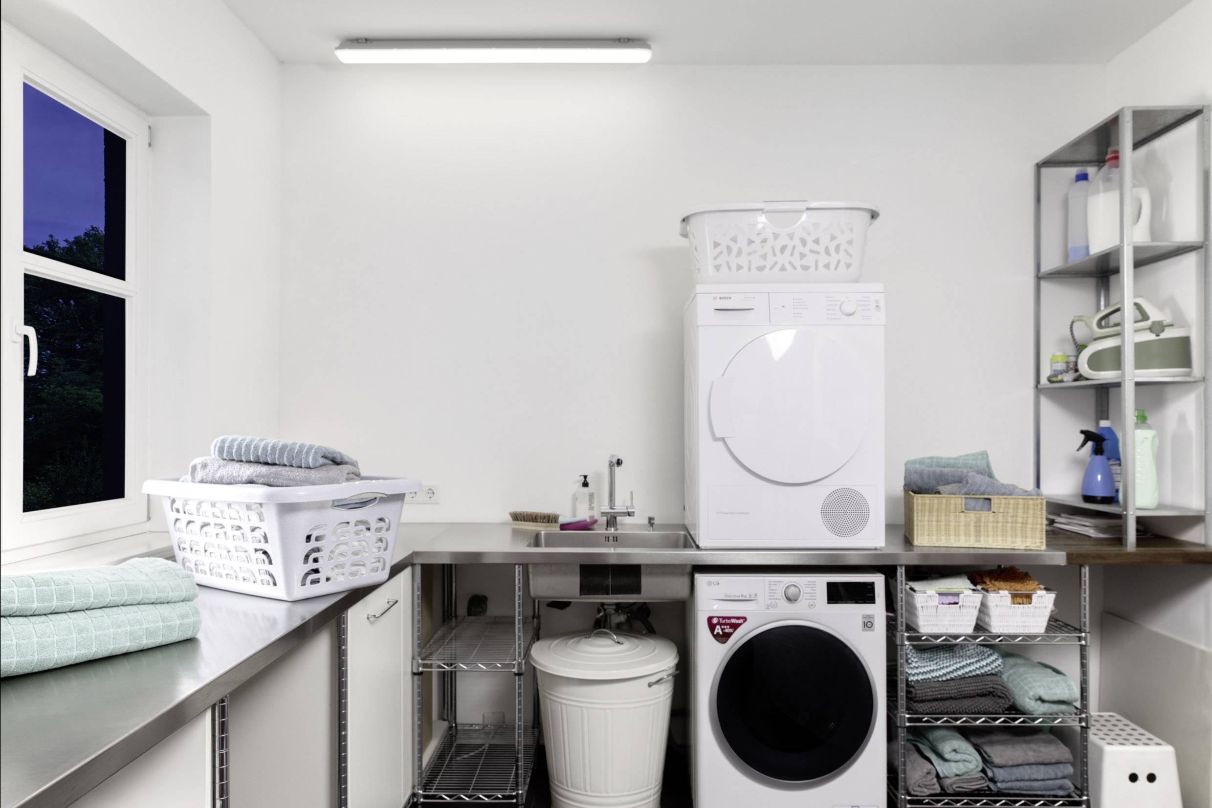 Ein moderner Hauswirtschaftsraum mit zwei Waschmaschinen, Wäschekörben auf den Maschinen, weißen Wänden und Regalen mit Putzmitteln.