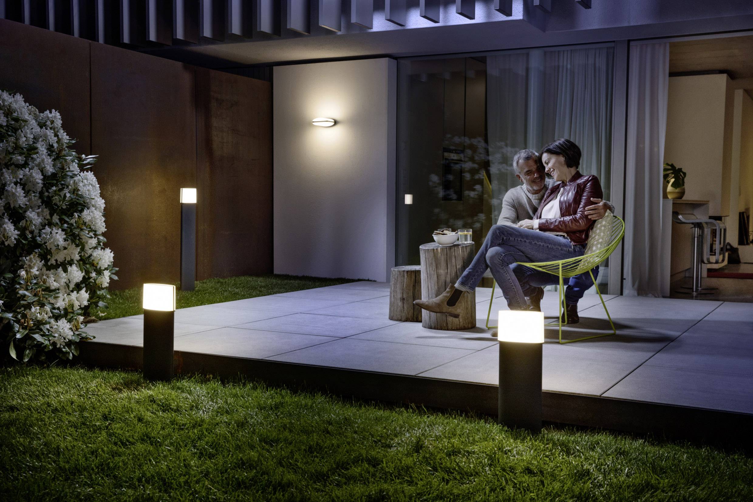 Ein Paar sitzt abends auf einer beleuchteten Terrasse, eingehüllt in Decken, umgeben von Blumen und leicht beleuchtetem Gras.