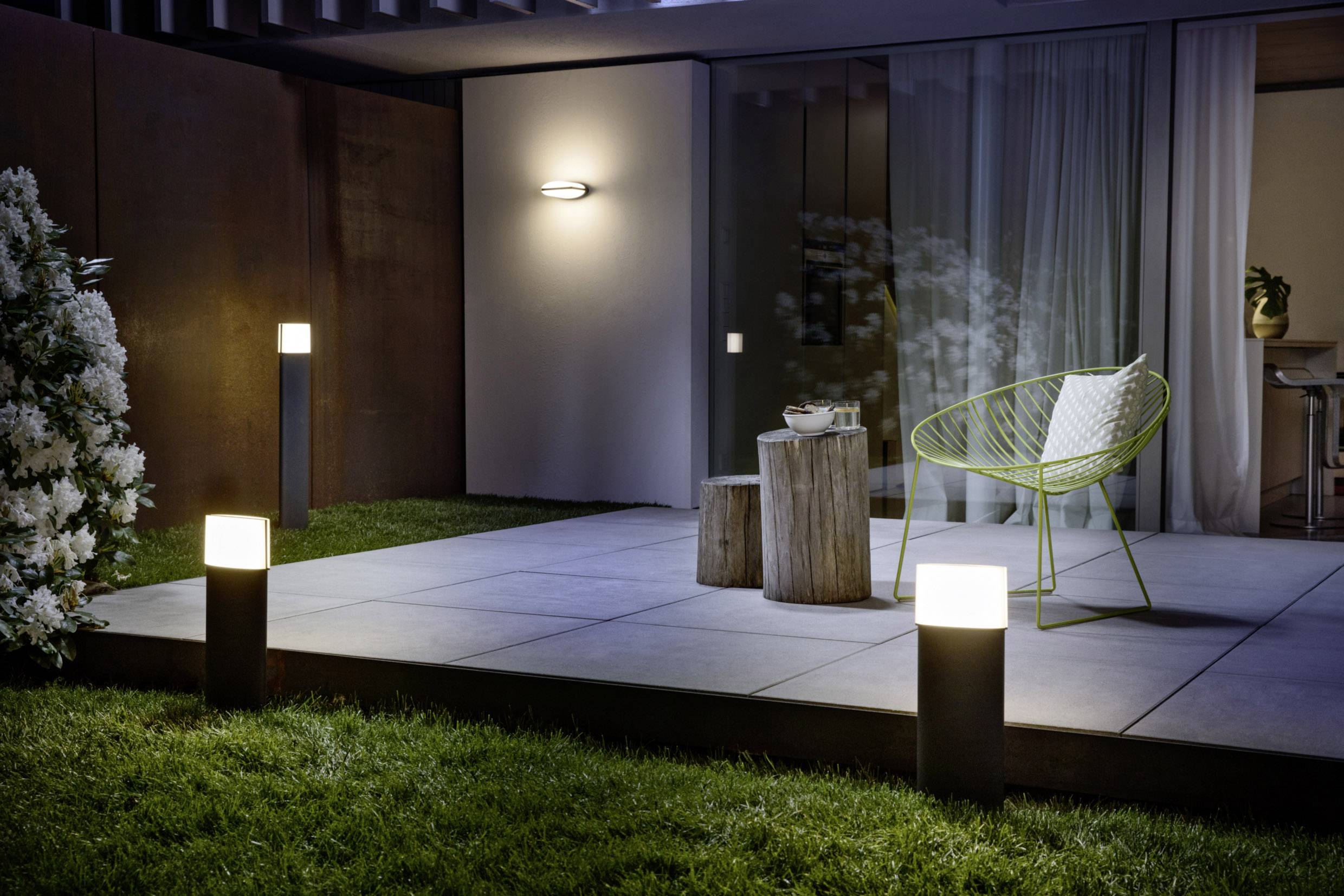 Eine moderne Terrasse bei Nacht mit Beleuchtung: drei Bodenlampen auf dem Rasen, ein grüner Stuhl, ein Tisch mit Dekor, und Pflanzen.