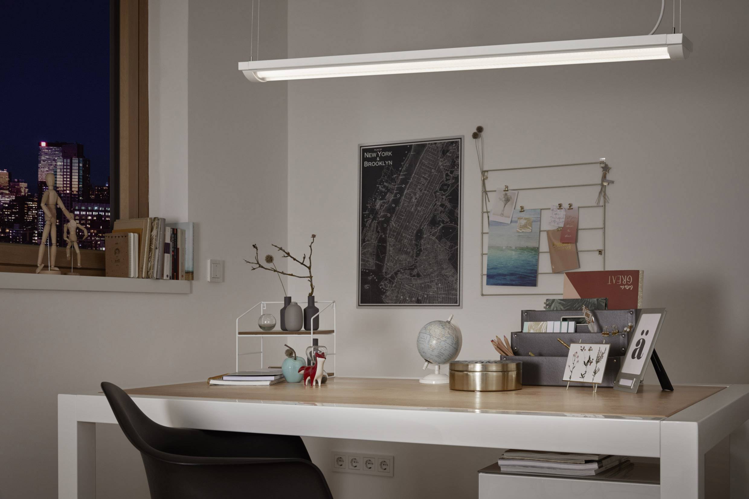 Ein Schreibtisch mit Lampe, Globus, Büchern und Dekofiguren vor einer Wand mit Bildern und einer Karte, im Hintergrund ein beleuchtetes Fenster.