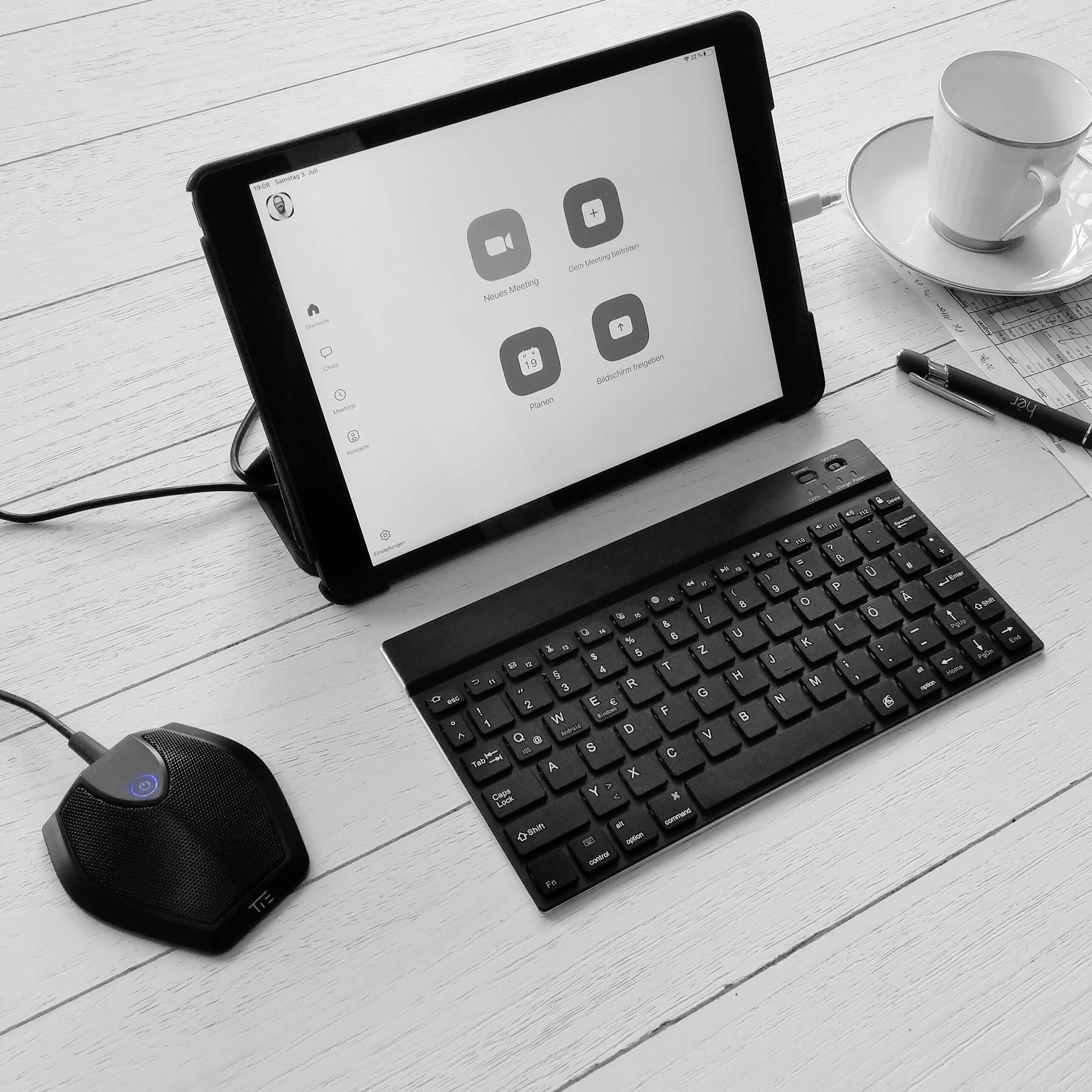 Tablet auf Tisch mit Videokonferenz-App geöffnet, daneben Tastatur und Kaffeetasse. Gerät bereit für Online-Meeting.