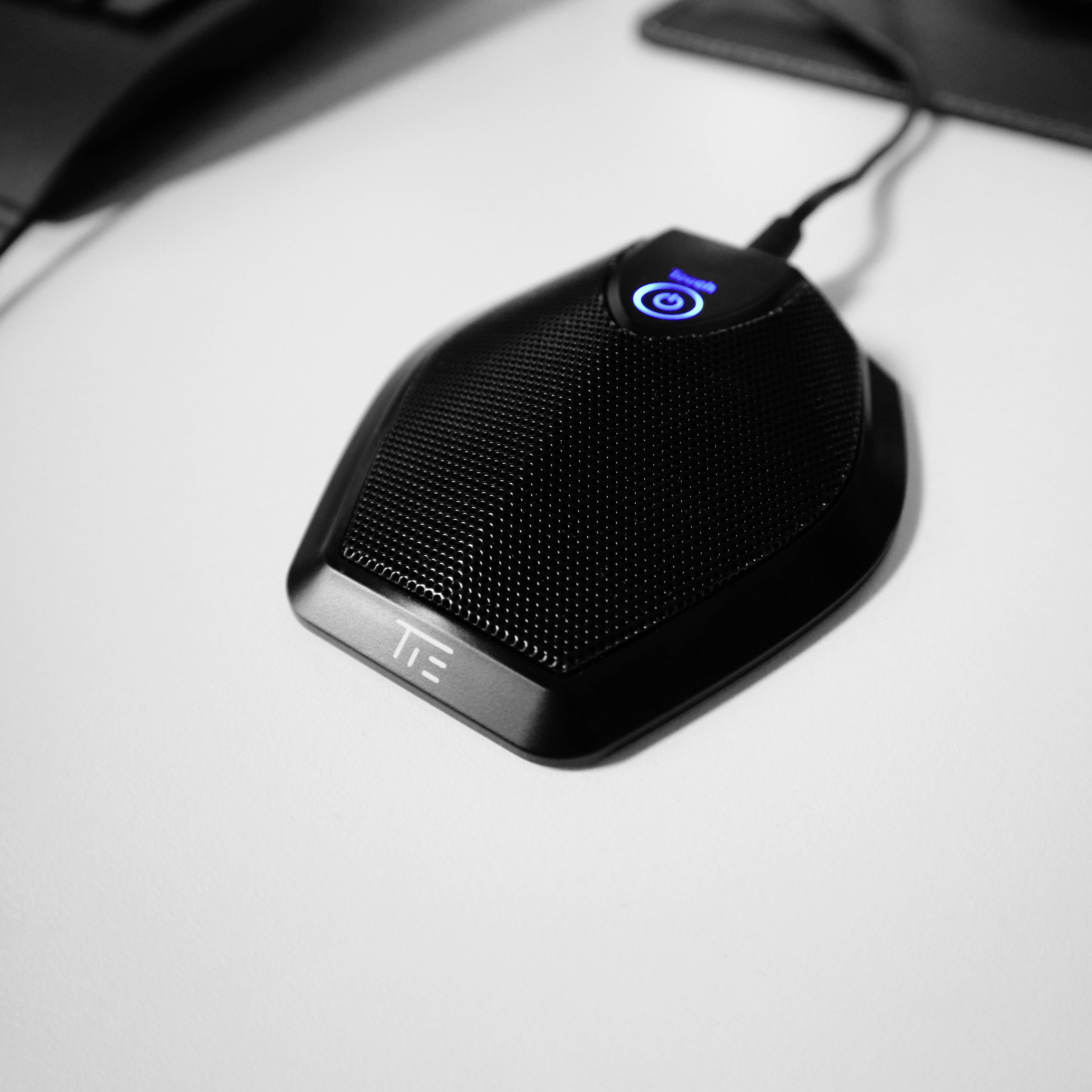 Ein schwarzes Tischmikrofon auf einem weißen Tisch, mit einem leuchtend blauen Einschaltknopf, das bereit für Sprachaufnahmen ist.