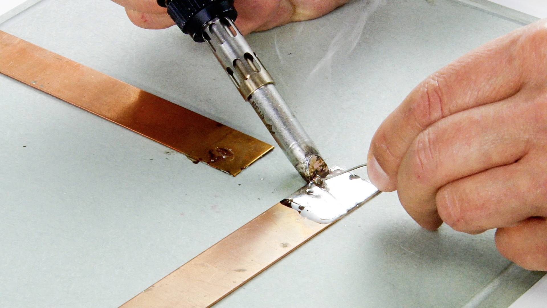 Eine Person lötet mit einem Lötkolben zwei Kupferstreifen auf einer glatten Oberfläche zusammen.
