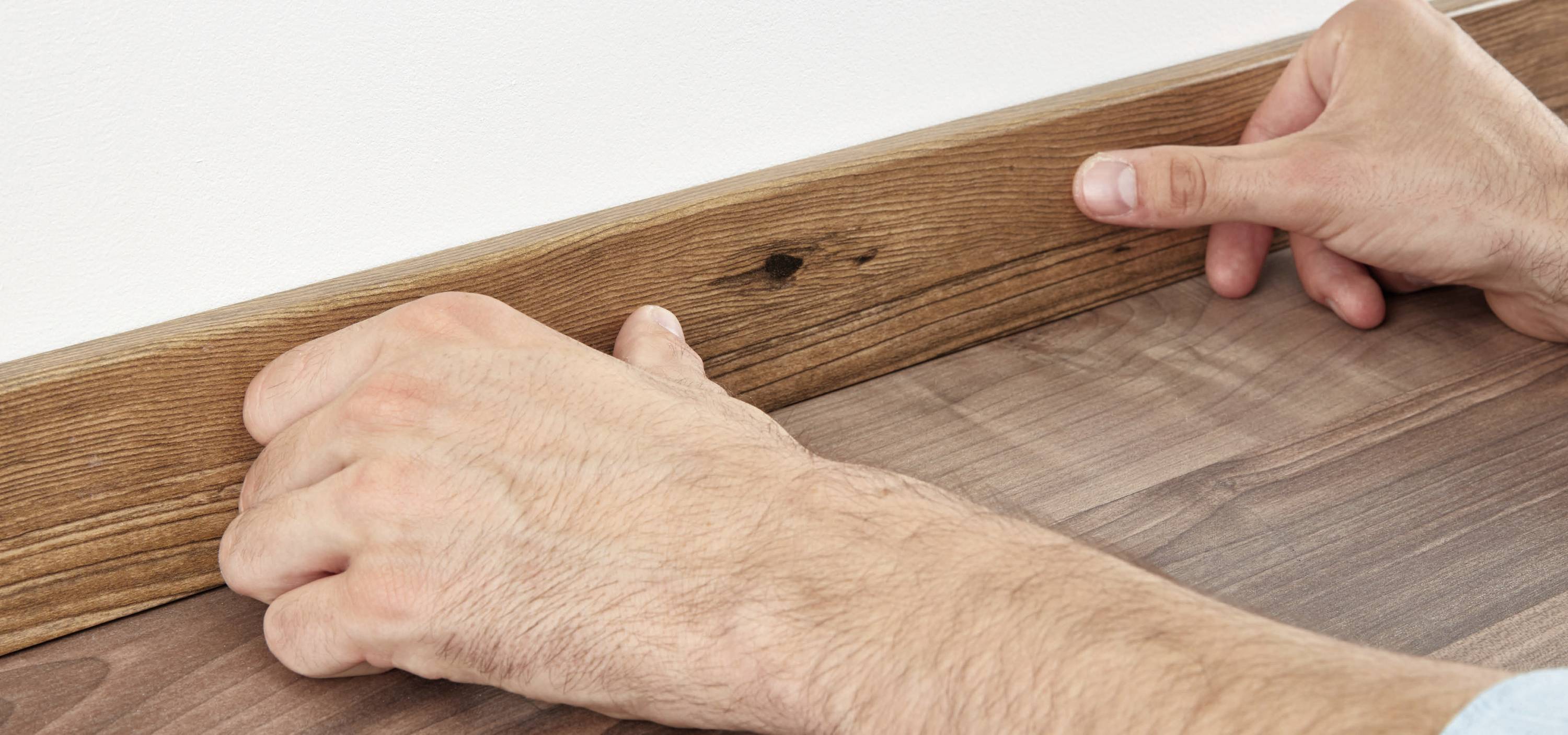 Zwei Hände installieren eine braune Holzleiste an einem Holzboden entlang einer weißen Wand.