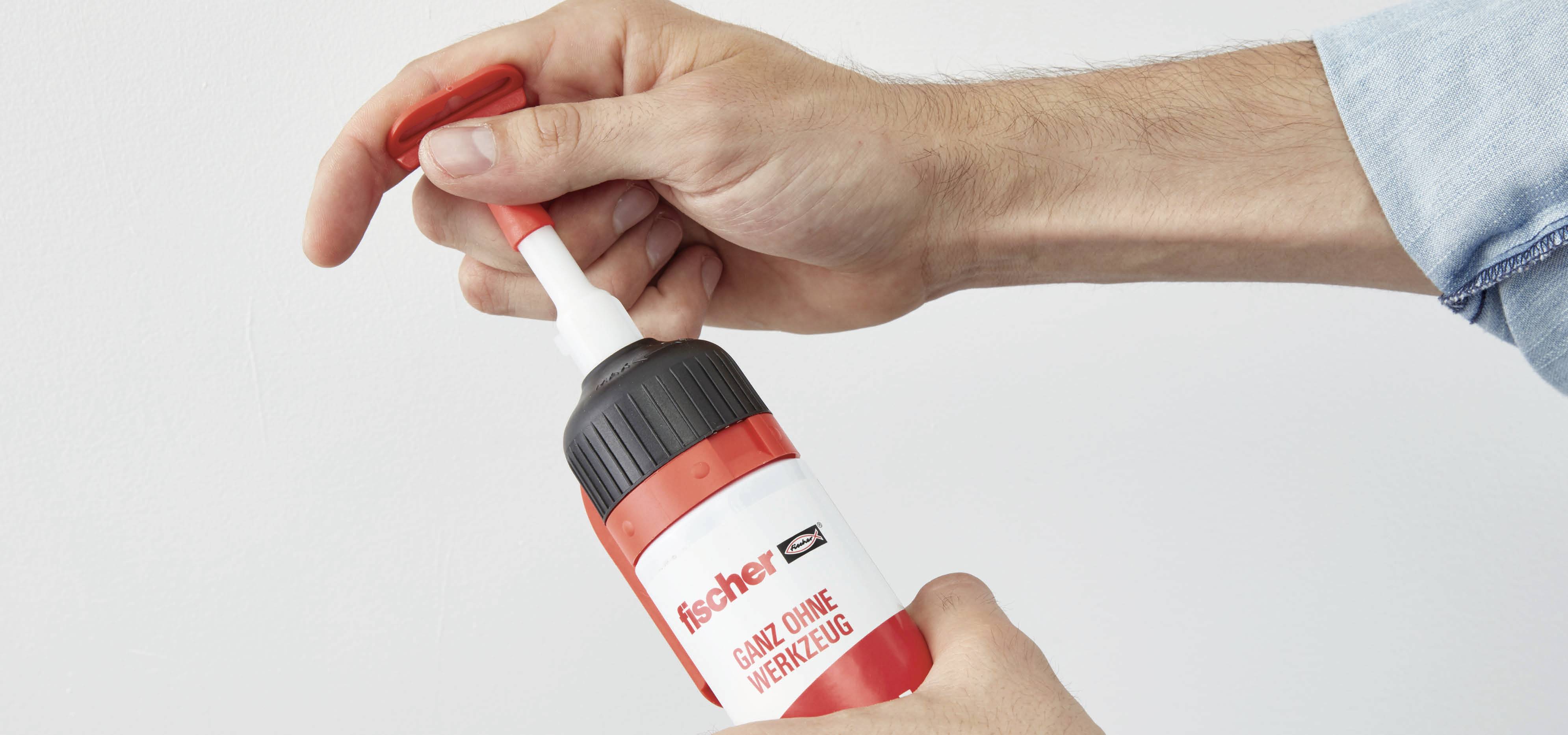 Eine Hand hält eine rote Klebetube von 'fischer', während die andere Hand die weiße Verschlusskappe entfernt.