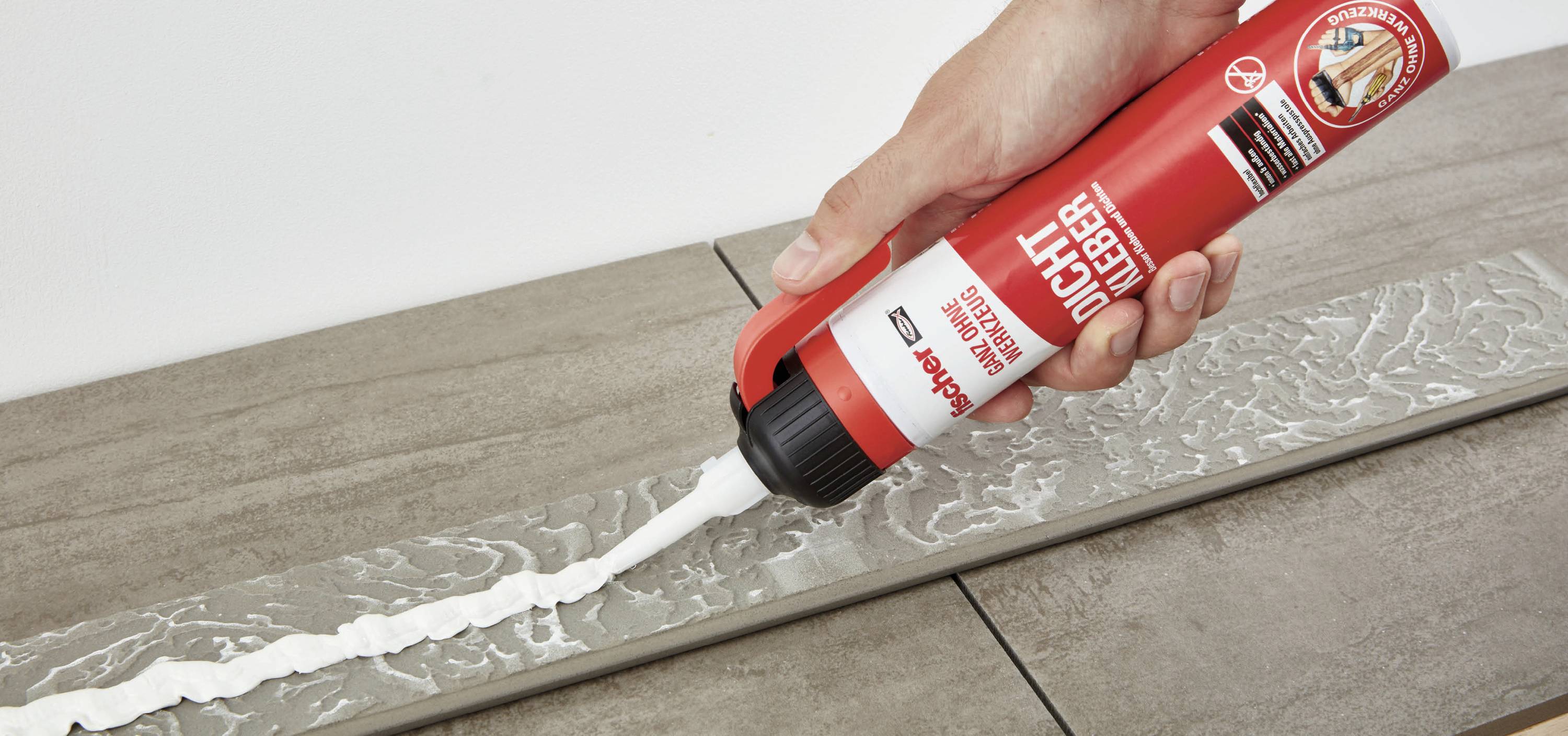 Eine Hand drückt weißen Kleber aus einer roten Tube auf eine graue Fliese.