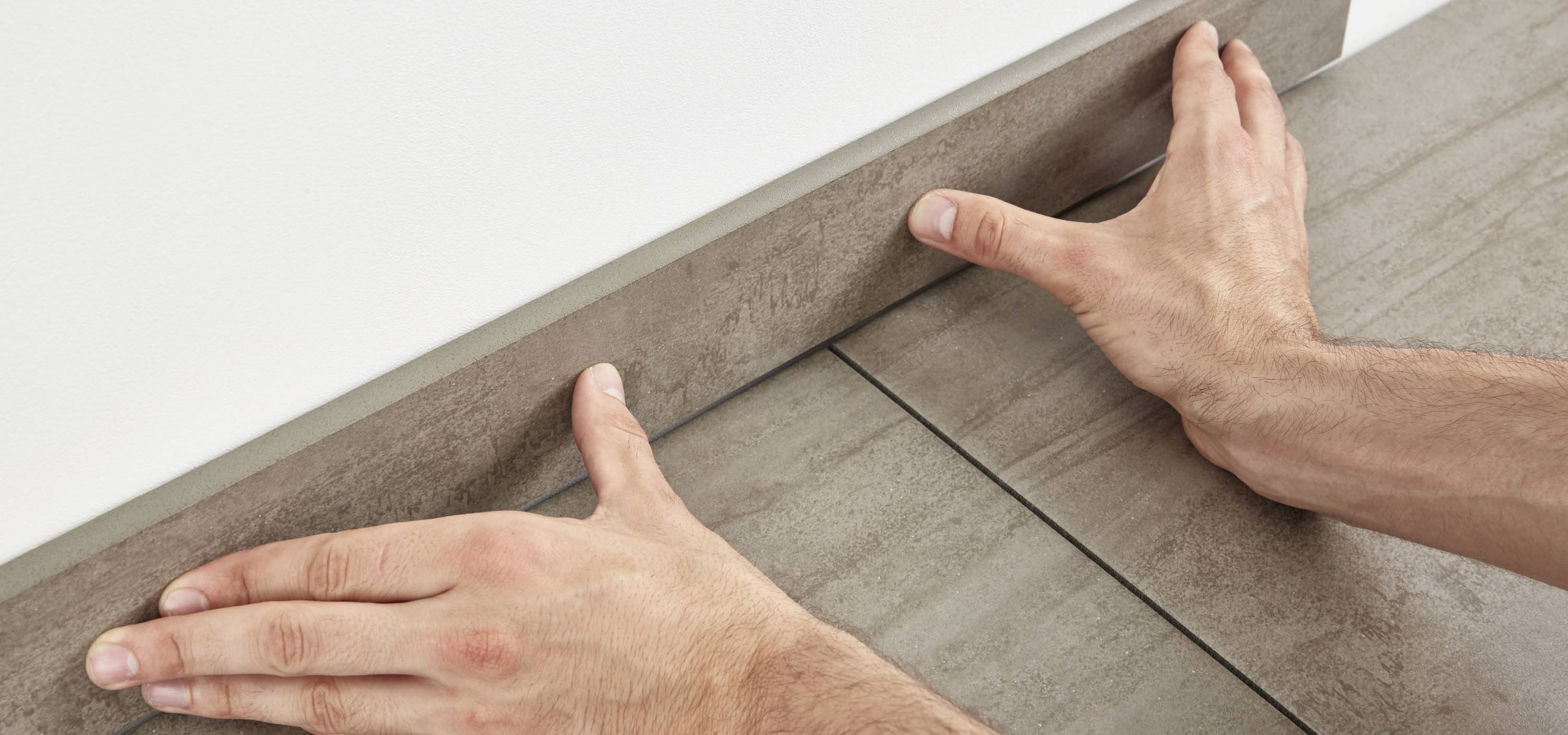 Zwei Hände setzen eine Fußleiste an eine Wand über grauen Fliesen ein und richten sie aus.