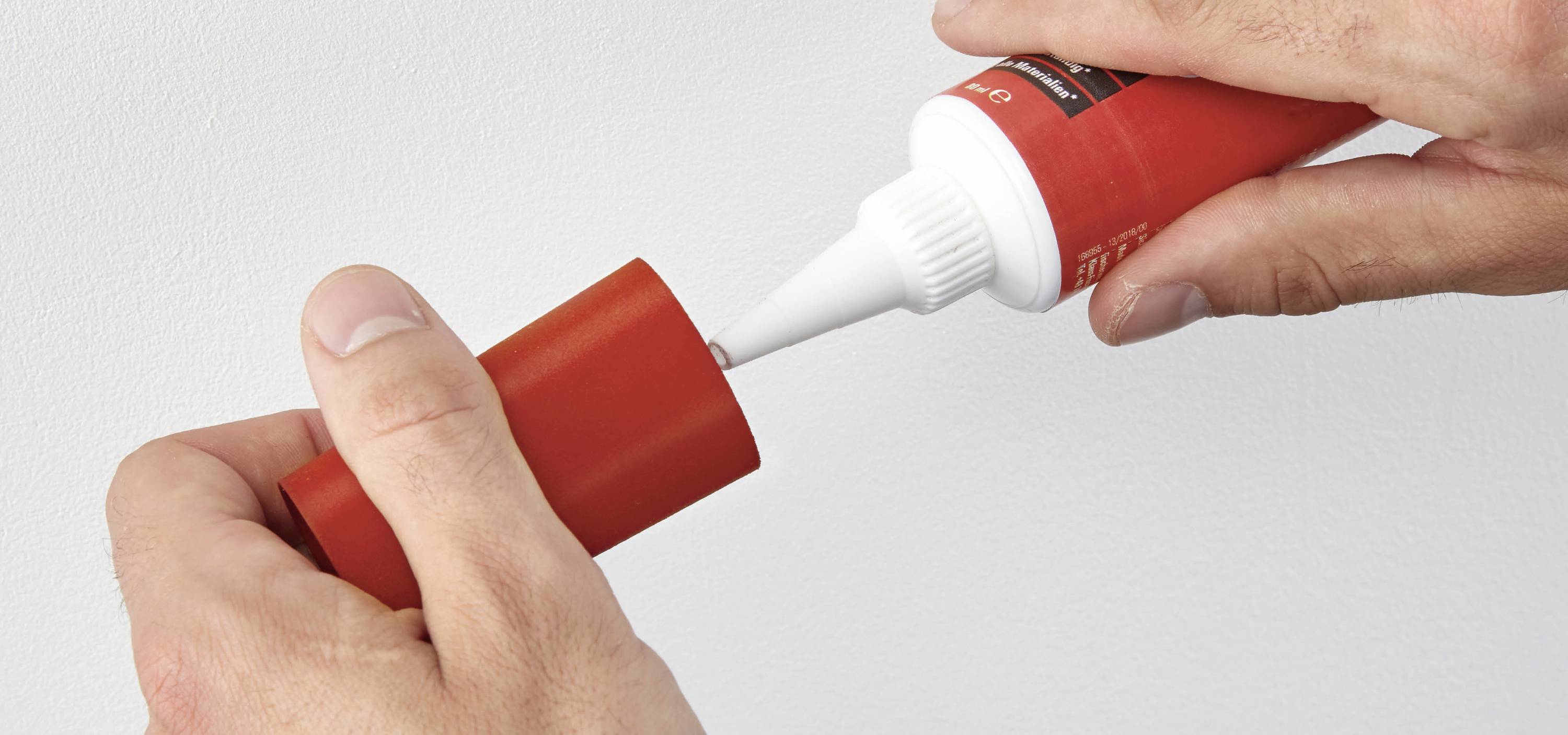 Eine Person verwendet eine rote Tube, um eine Substanz auf eine rote Kappe aufzutragen. Hände zeigen klar den Vorgang.