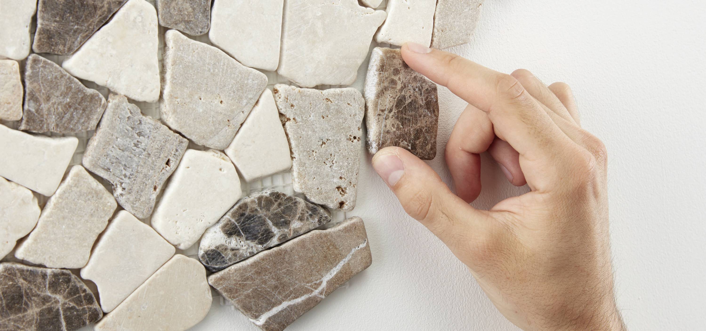 Eine Hand fügt ein braunes Mosaikstück in ein Wandmosaik ein. Die Mosaiksteine sind in verschiedenen Erdtönen gehalten.