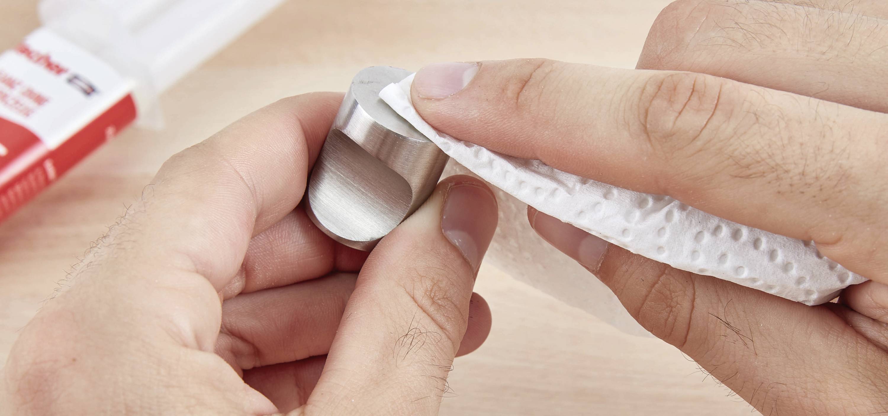 Eine Hand hält einen Metallzylinder, während die andere die Oberfläche mit einem Tuch reinigt. Im Hintergrund ist Kleber zu sehen.