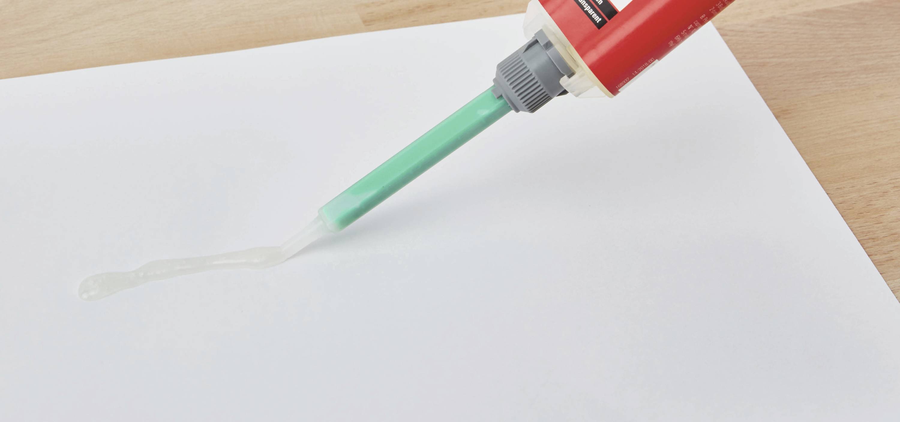 Eine rote Klebeflasche liegt auf einem Tisch und gibt einen langen Streifen Klebstoff auf ein weißes Papier ab.