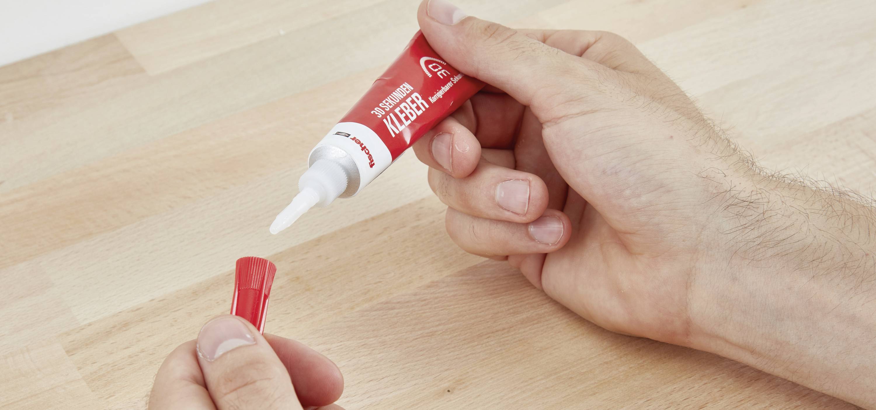 Eine Hand hält eine rote Tube Klebstoff über einem Holztisch, während die andere Hand die Kappe entfernt.