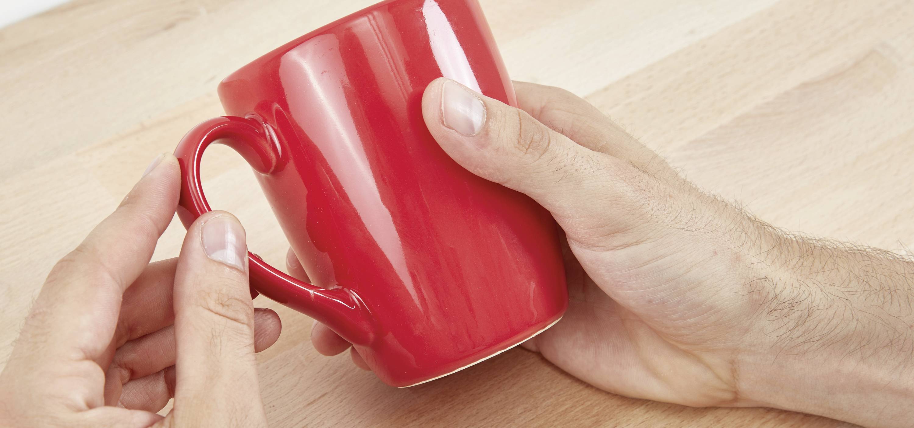 Zwei Hände halten eine rote Tasse auf einem Holztisch.