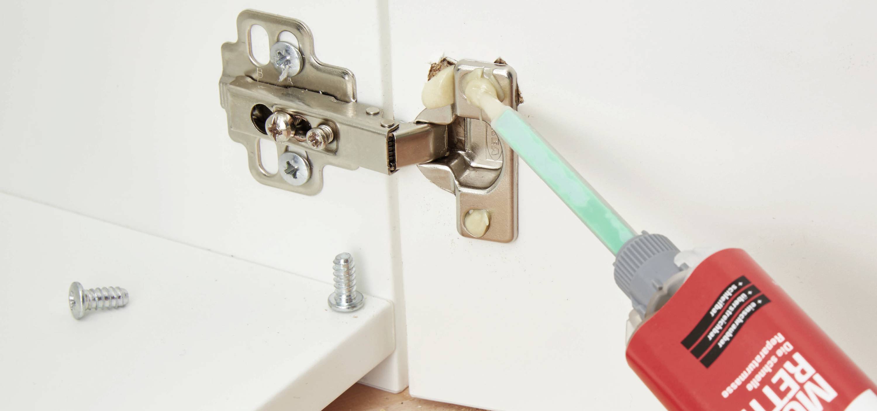 Eine Person klebt ein Scharnier an einem Schrank mit einem Kleber aus einer roten Tube. Zwei Schrauben liegen daneben bereit.