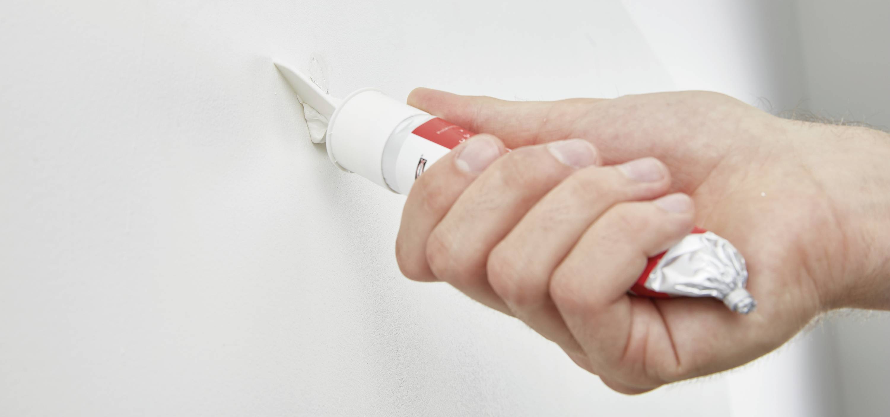'Hand drückt Spachtelmasse aus einer Tube in ein Loch in der Wand.'