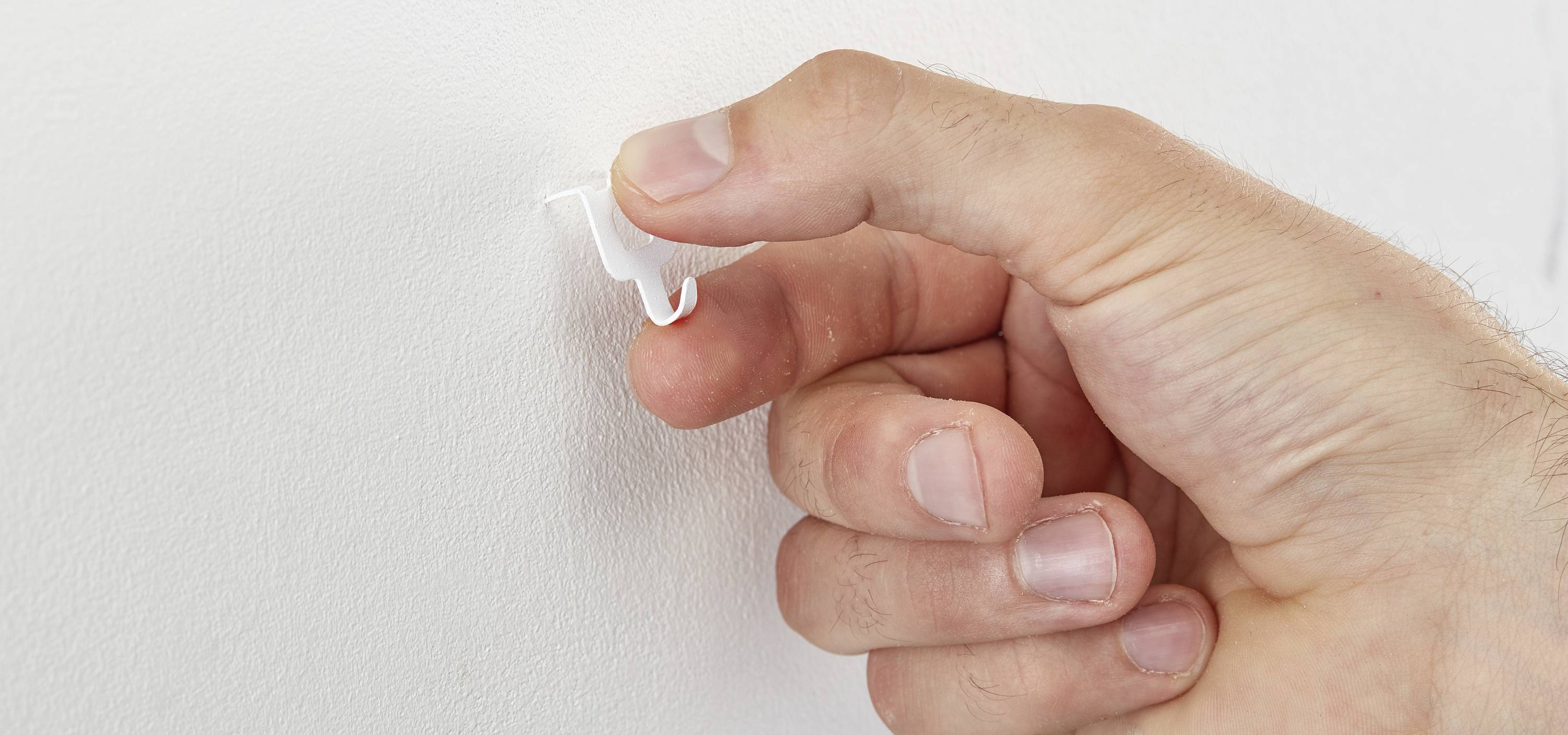 Eine Hand drückt einen kleinen Kunststoffhaken in eine weiße Wand. Der Haken wird benutzt, um leichte Gegenstände aufzuhängen.