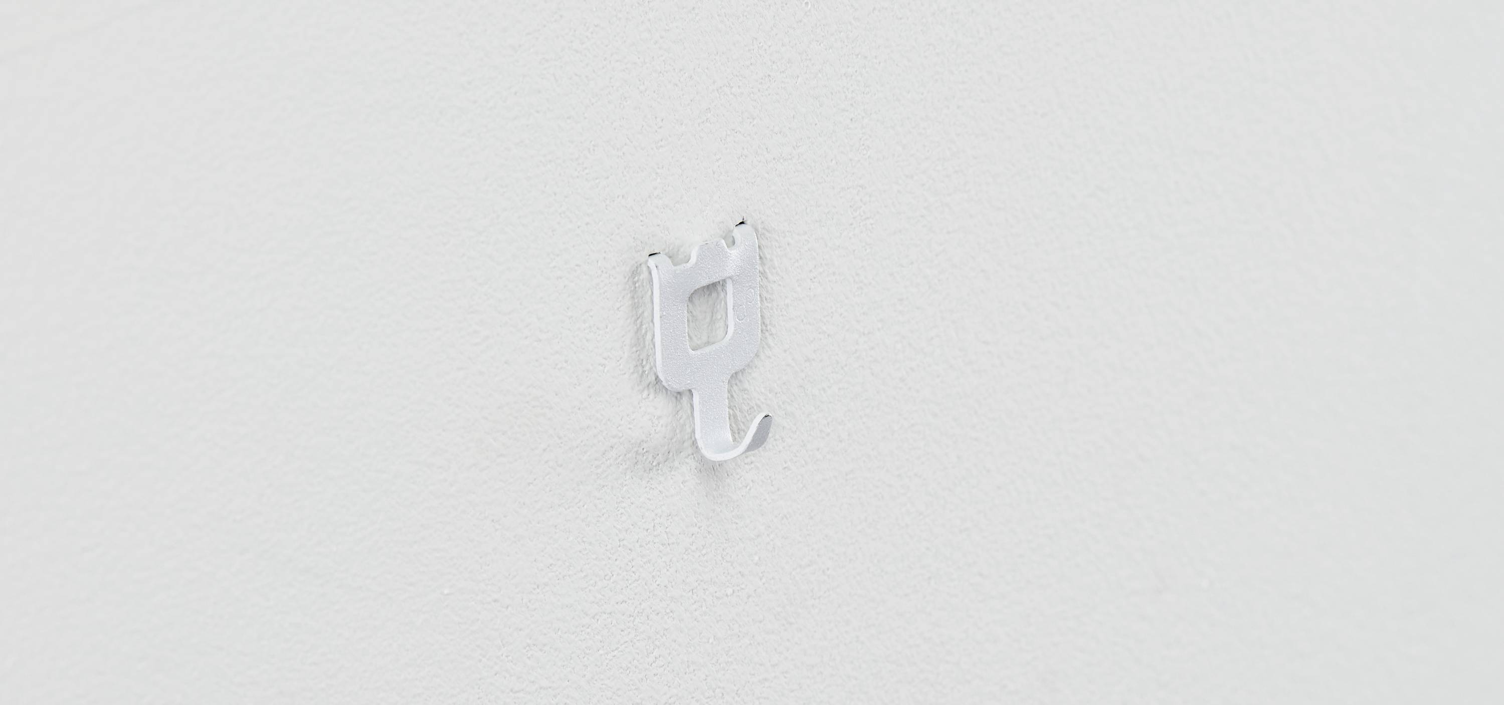 Eine kleine, metallische Wandhalterung in Form eines stilisierten Hakens auf einer weißen Oberfläche.