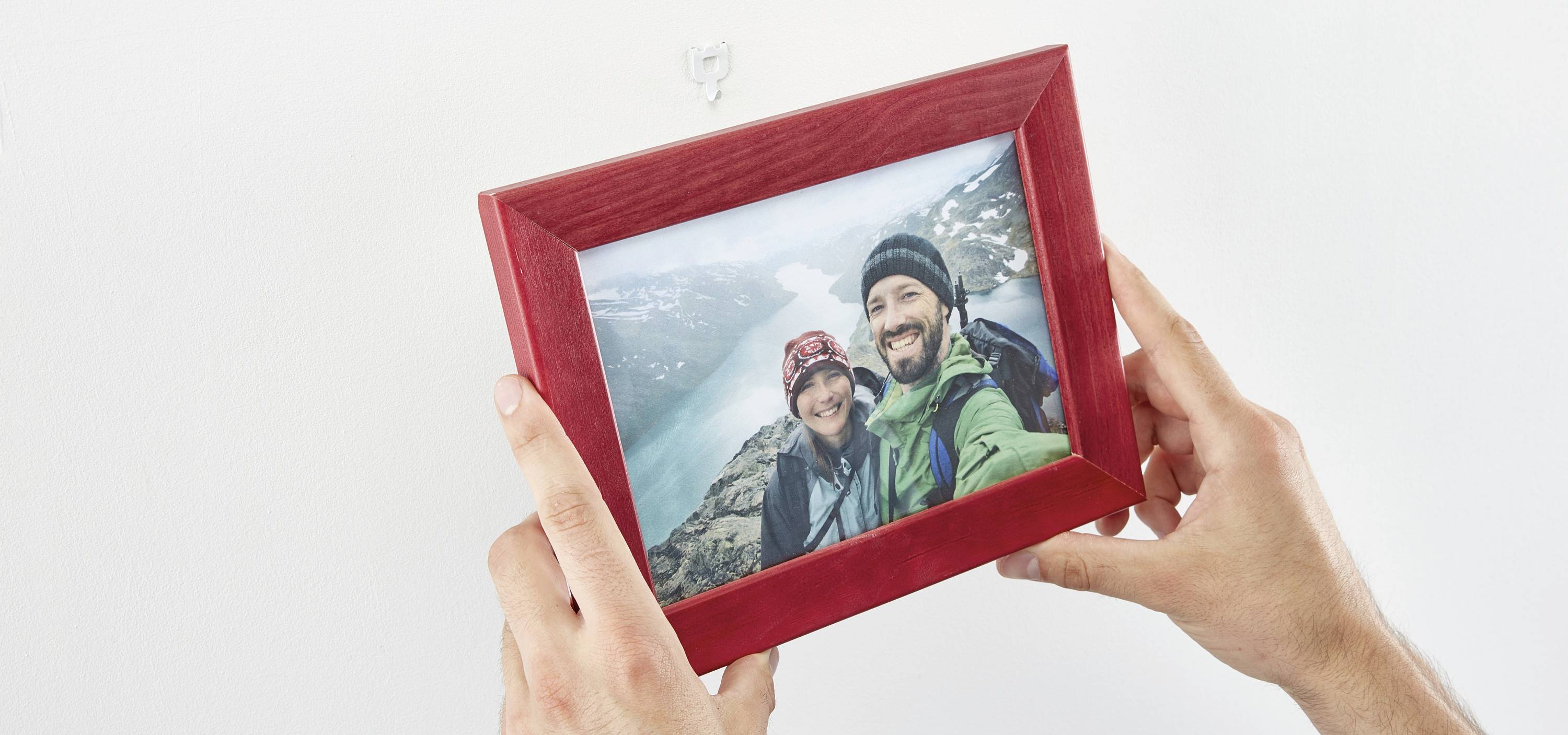 Person hält einen rot gerahmten Fotoabdruck von zwei lächelnden Menschen auf einer Bergwanderung mit See im Hintergrund.