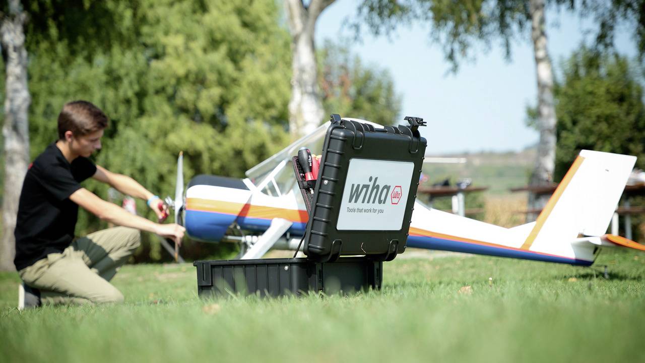 Ein Mann kniet auf einer Wiese und arbeitet an einem großen Modellflugzeug, neben ihm steht ein schwarzer Werkzeugkoffer von Wiha.