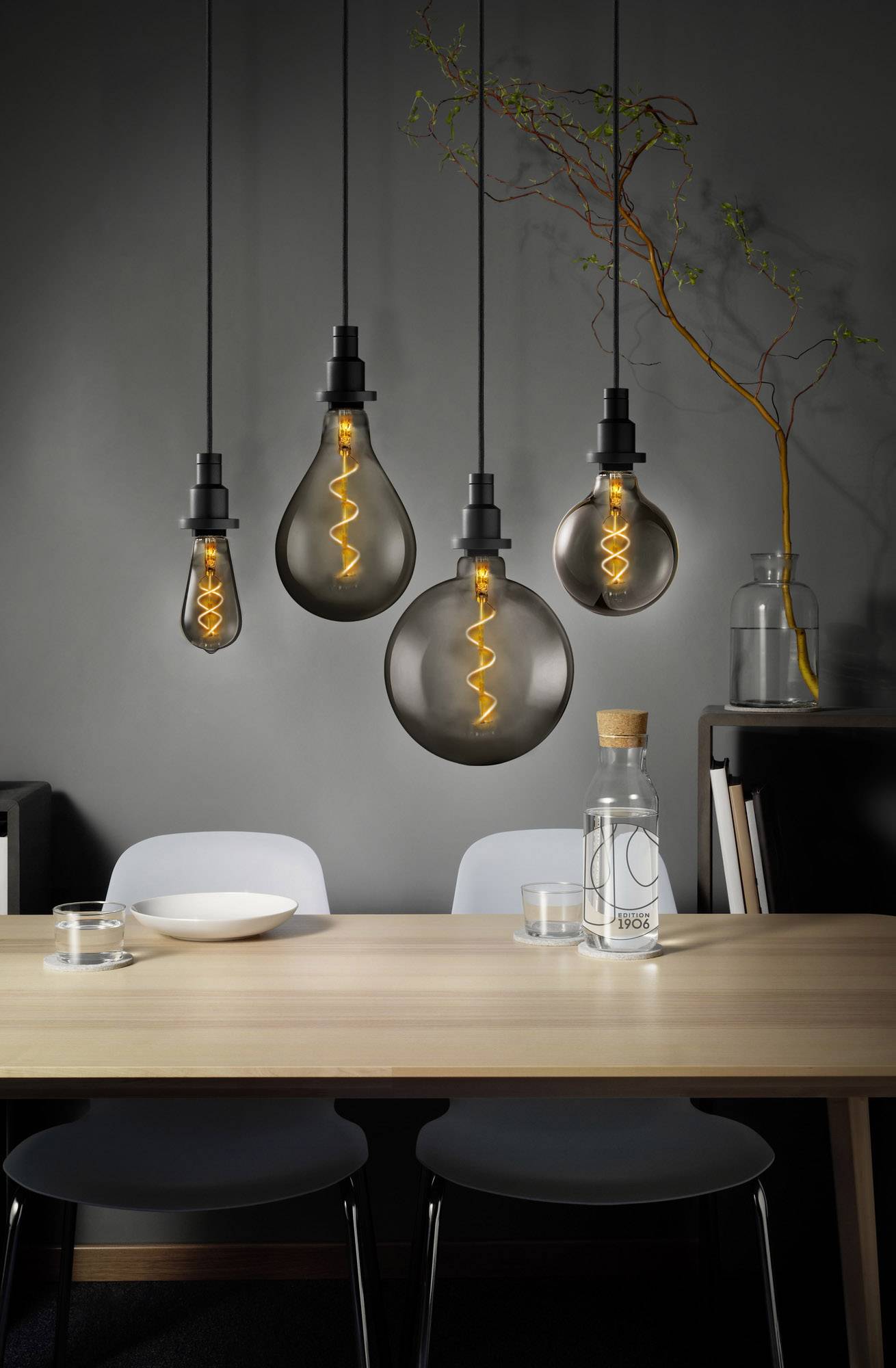 Ein modernes Esszimmer mit Holz­tisch, darauf ein Wasserkaraffe und Gläser. Oben hängen stilvolle Glühbirnen mit leuchtenden Filamenten.