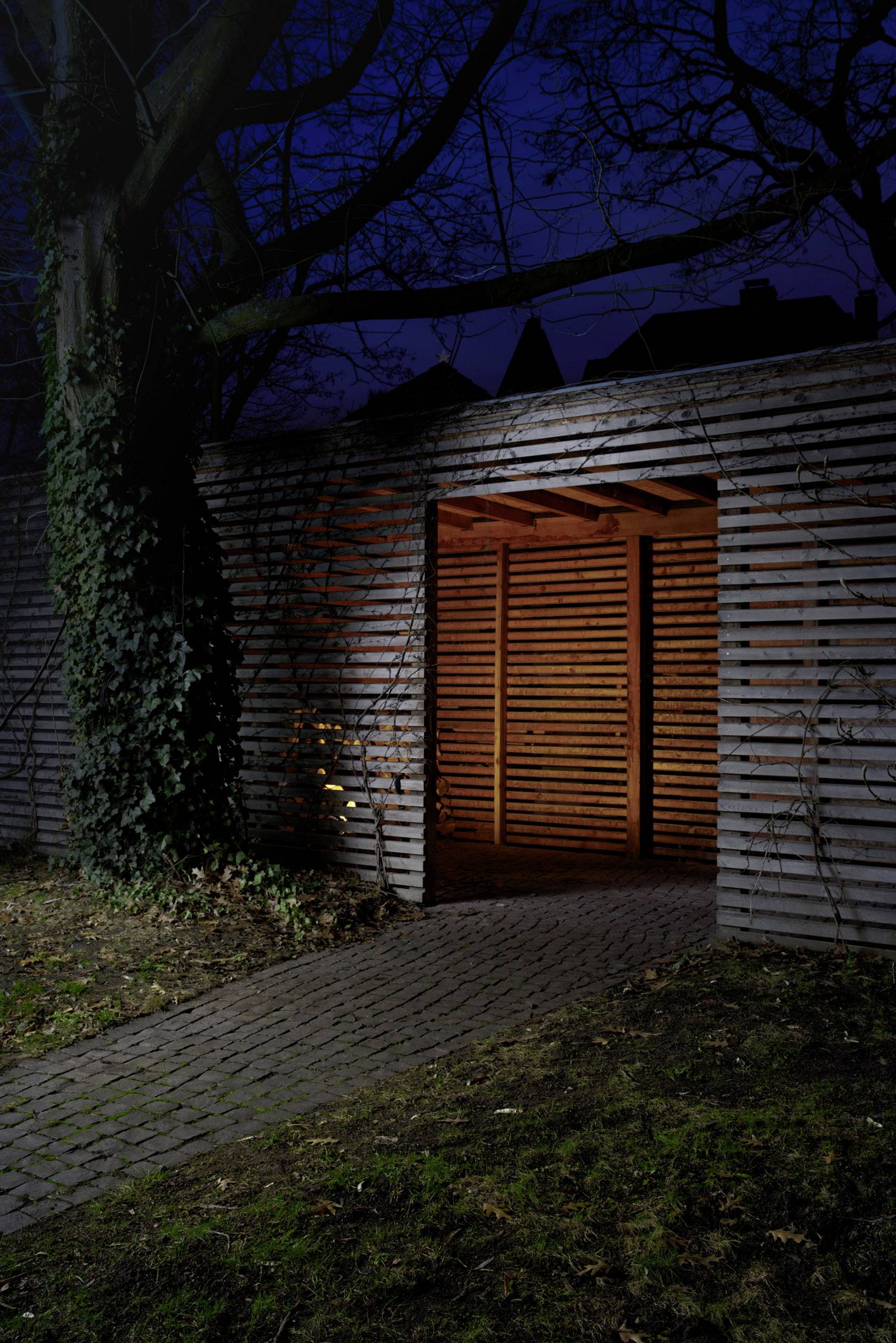 Holzpergola bei Nacht, beleuchtet von innen. Ein angeleuchteter Baum steht links, Weg führt zur Pergola.