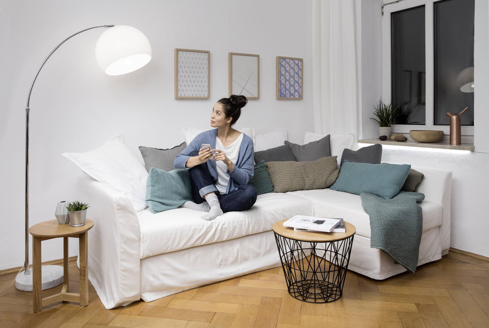 Eine Frau sitzt auf einem weißen Sofa in einem gemütlich eingerichteten Wohnzimmer. Sie hält eine Tasse und schaut lächelnd zur Seite.