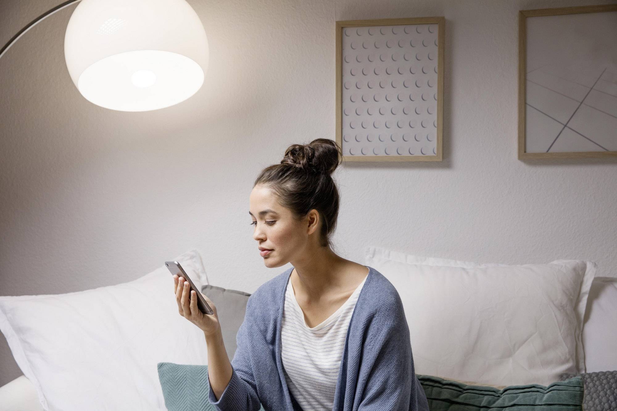 Frau sitzt auf dem Sofa und blickt auf ihr Smartphone. Sie trägt einen blauen Schal. Eine Lampe leuchtet hell im Hintergrund.