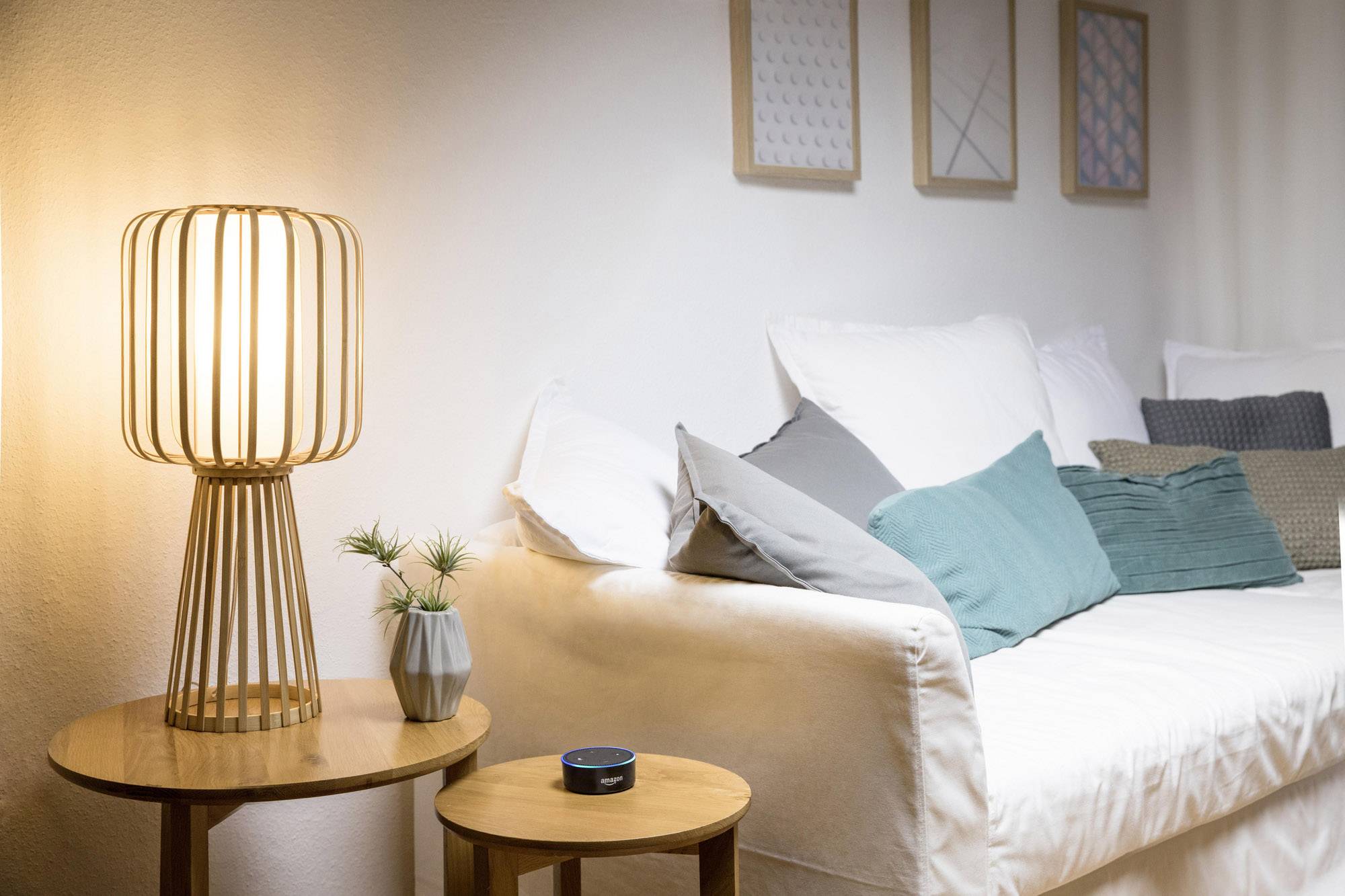 Ein gemütliches Wohnzimmer mit einem Sofa voller Kissen. Links im Vordergrund steht eine Tischlampe auf einem Holztisch, daneben ein kleiner Lautsprecher.