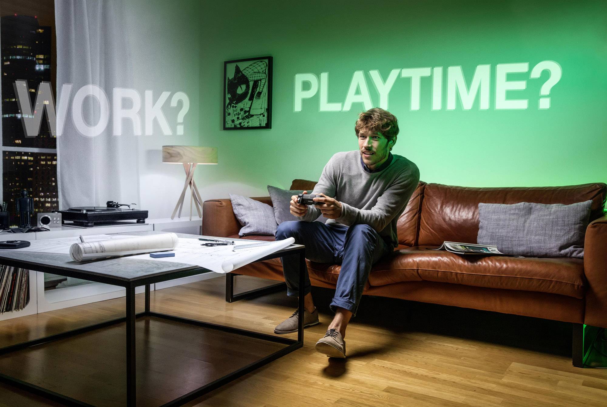 Ein Mann sitzt auf einem Sofa und spielt ein Videospiel. Hinter ihm sind die projizierten Worte 'WORK?' und 'PLAYTIME?' an der Wand zu sehen.