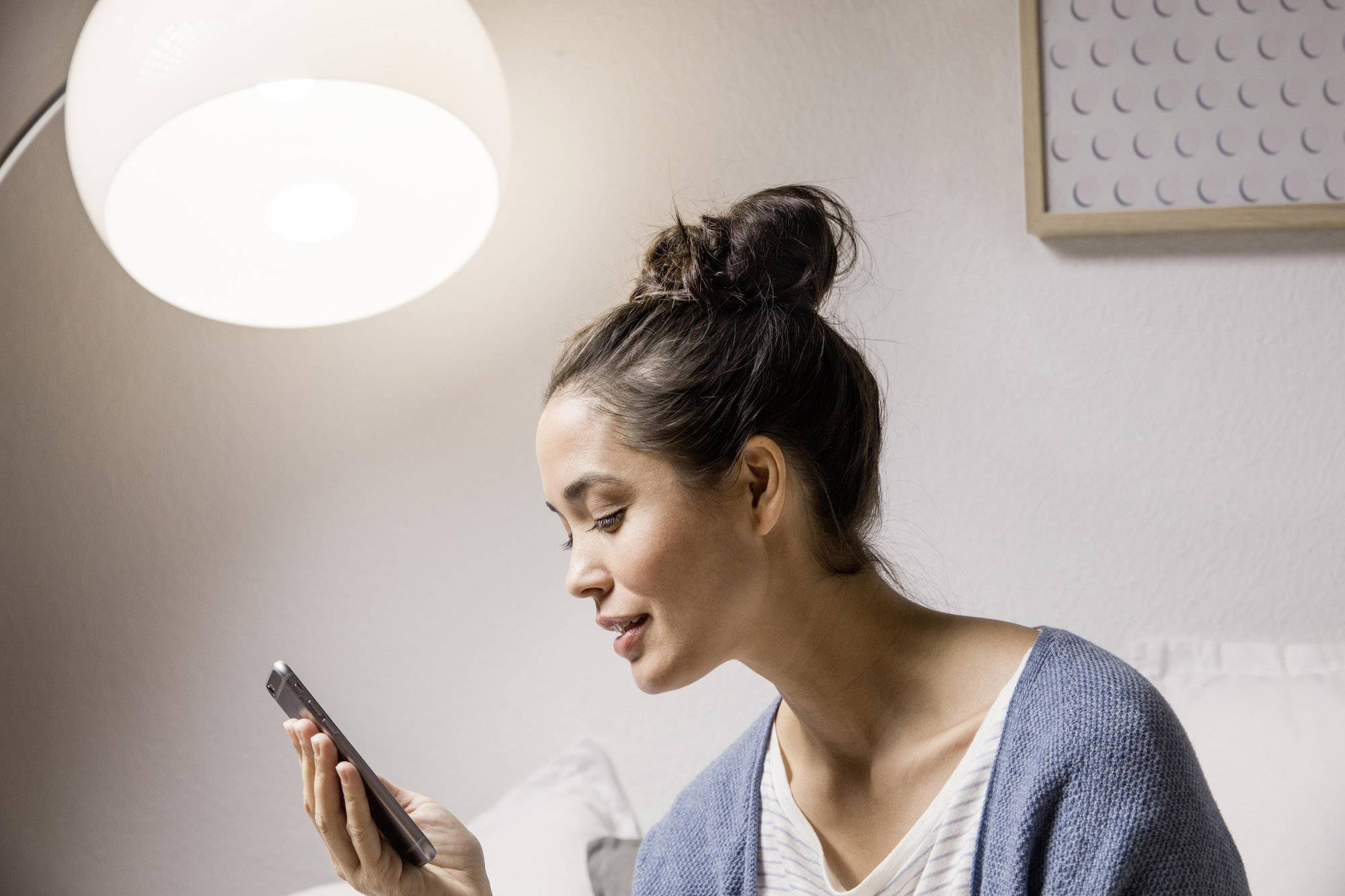 Eine Frau sitzt auf einem Sofa in einem gemütlichen Raum und spricht in ihr Smartphone. Eine Lampe leuchtet im Hintergrund.