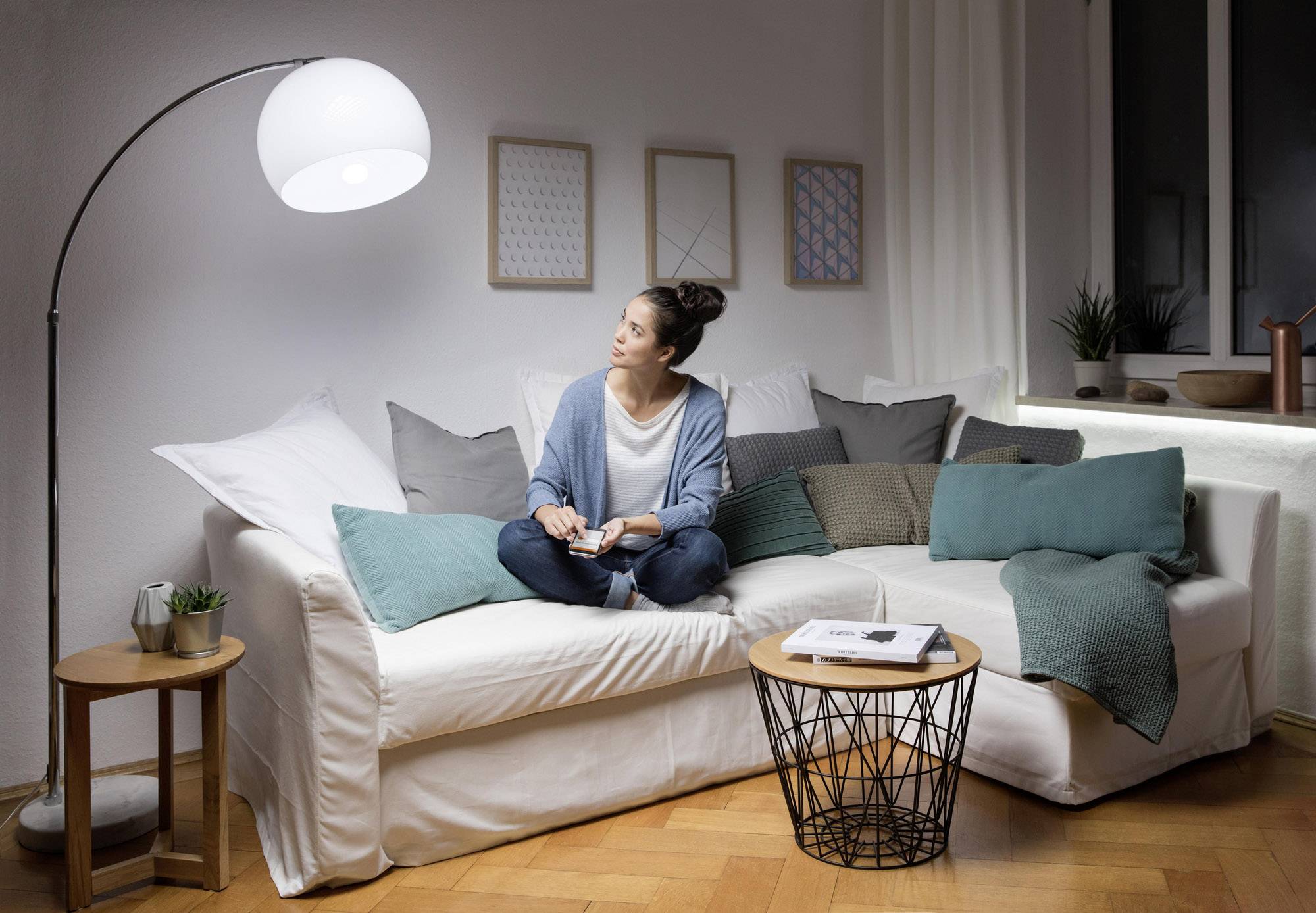 Eine Frau sitzt entspannt auf einem weißen Sofa in einem Wohnzimmer, umgeben von Kissen, und liest ein Buch bei gedämpftem Lampenlicht.