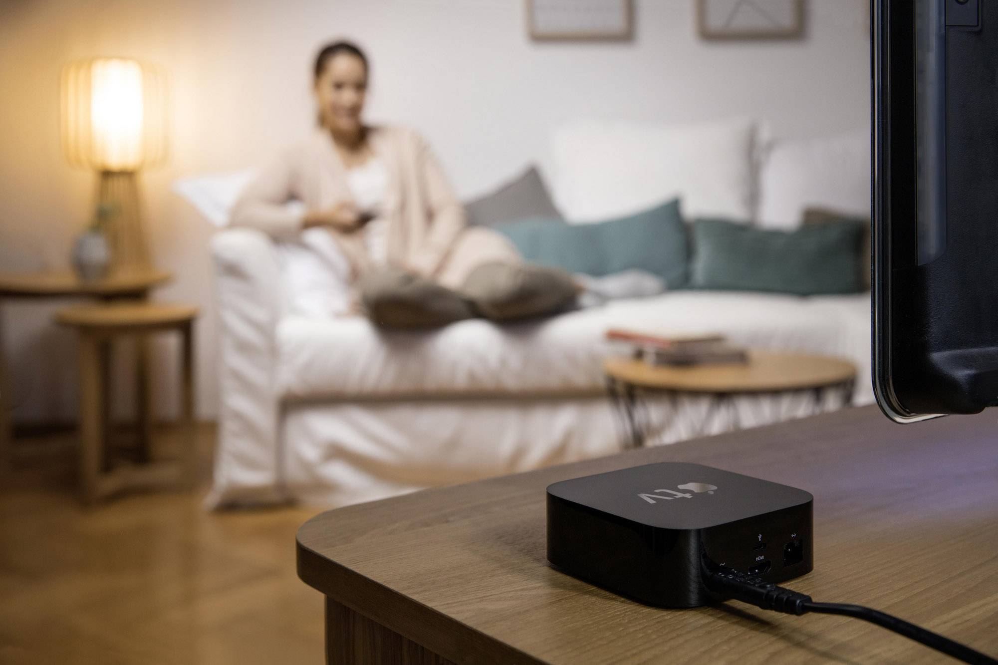 Eine Person sitzt entspannt auf einem Sofa im Hintergrund, während im Vordergrund ein Streaming-Gerät nahe einem Fernseher steht.