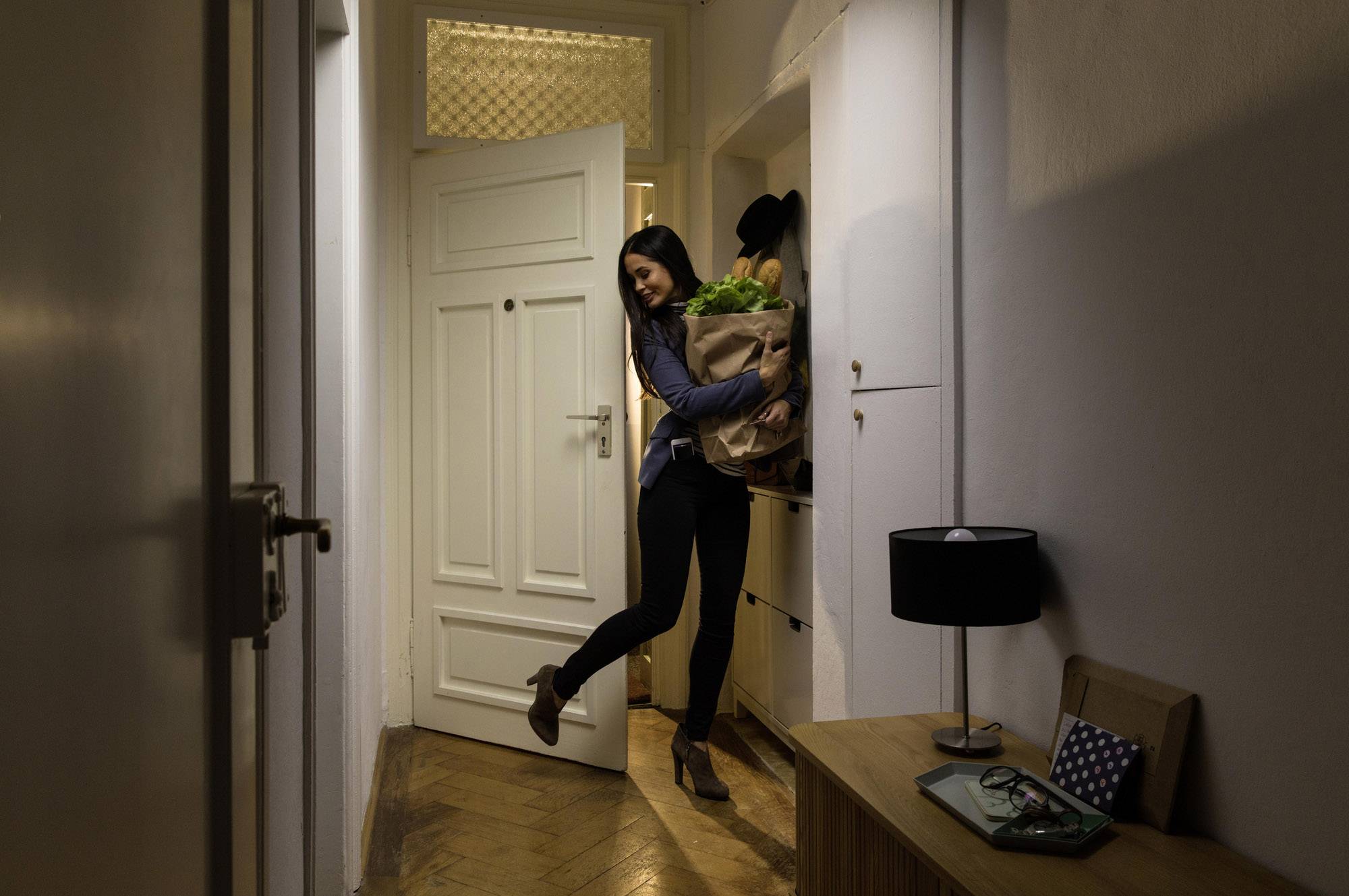 Eine Frau betritt eine Wohnung und hält eine Tasche mit Lebensmitteln in den Armen. Die Tür ist geöffnet, und es ist Abend.