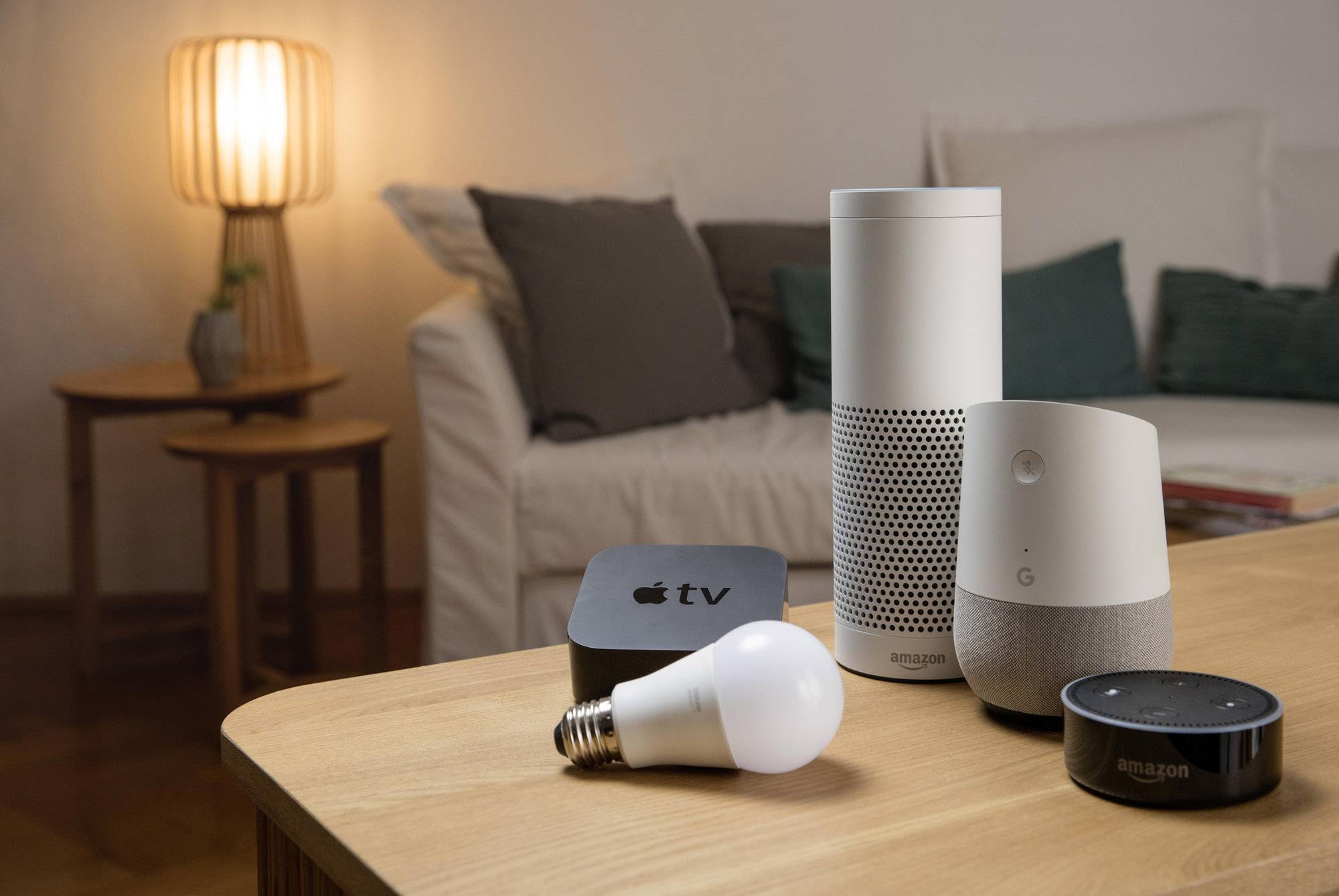 Smart-Home-Geräte auf einem Tisch in einem Wohnzimmer: Lautsprecher, Glühbirne, TV-Box. Im Hintergrund ein beleuchtetes Sofa.