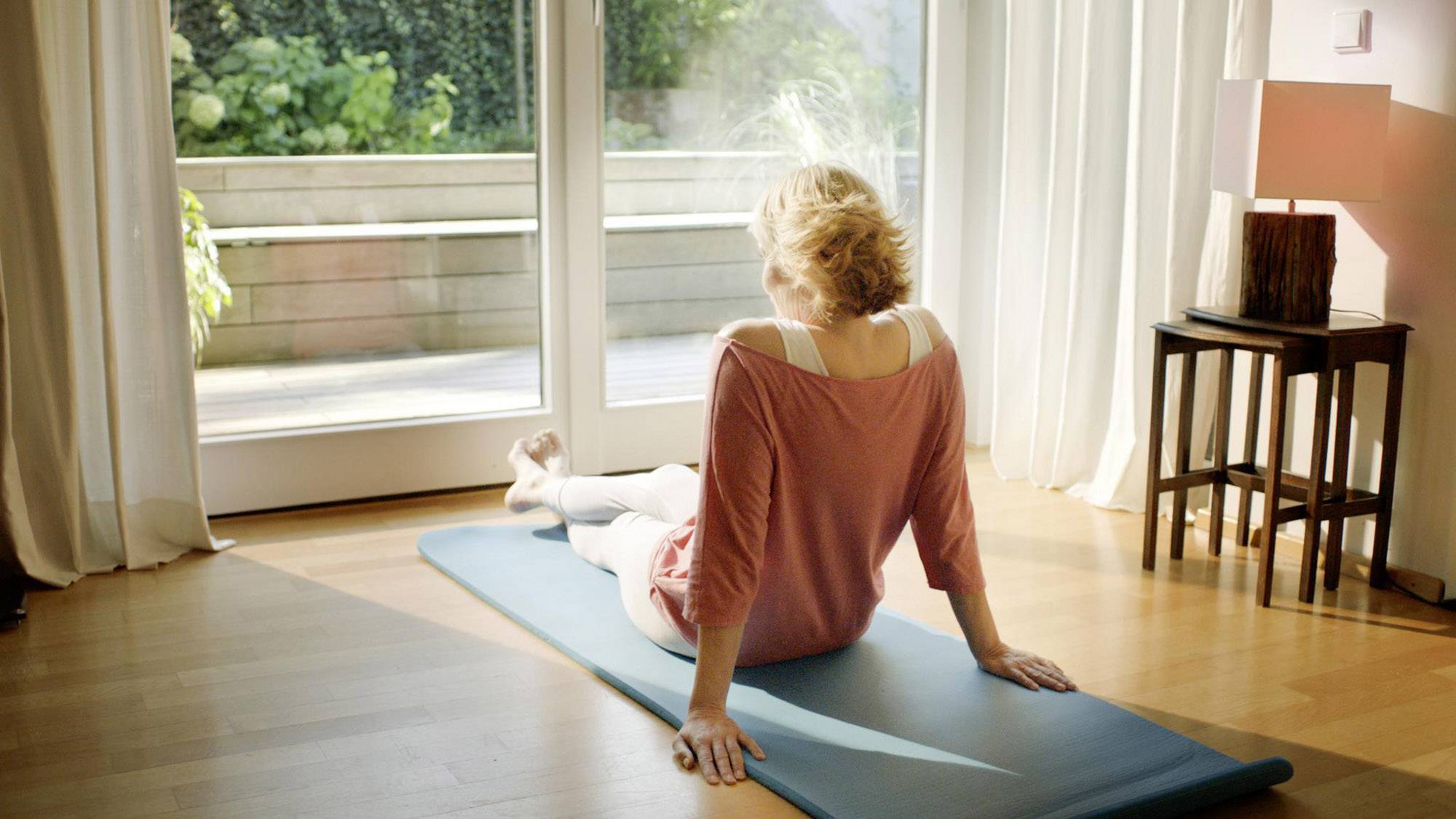 Eine Frau macht Dehnübungen auf einer Yogamatte in einem hellen Raum vor einer großen Fensterfront mit Blick ins Grüne.