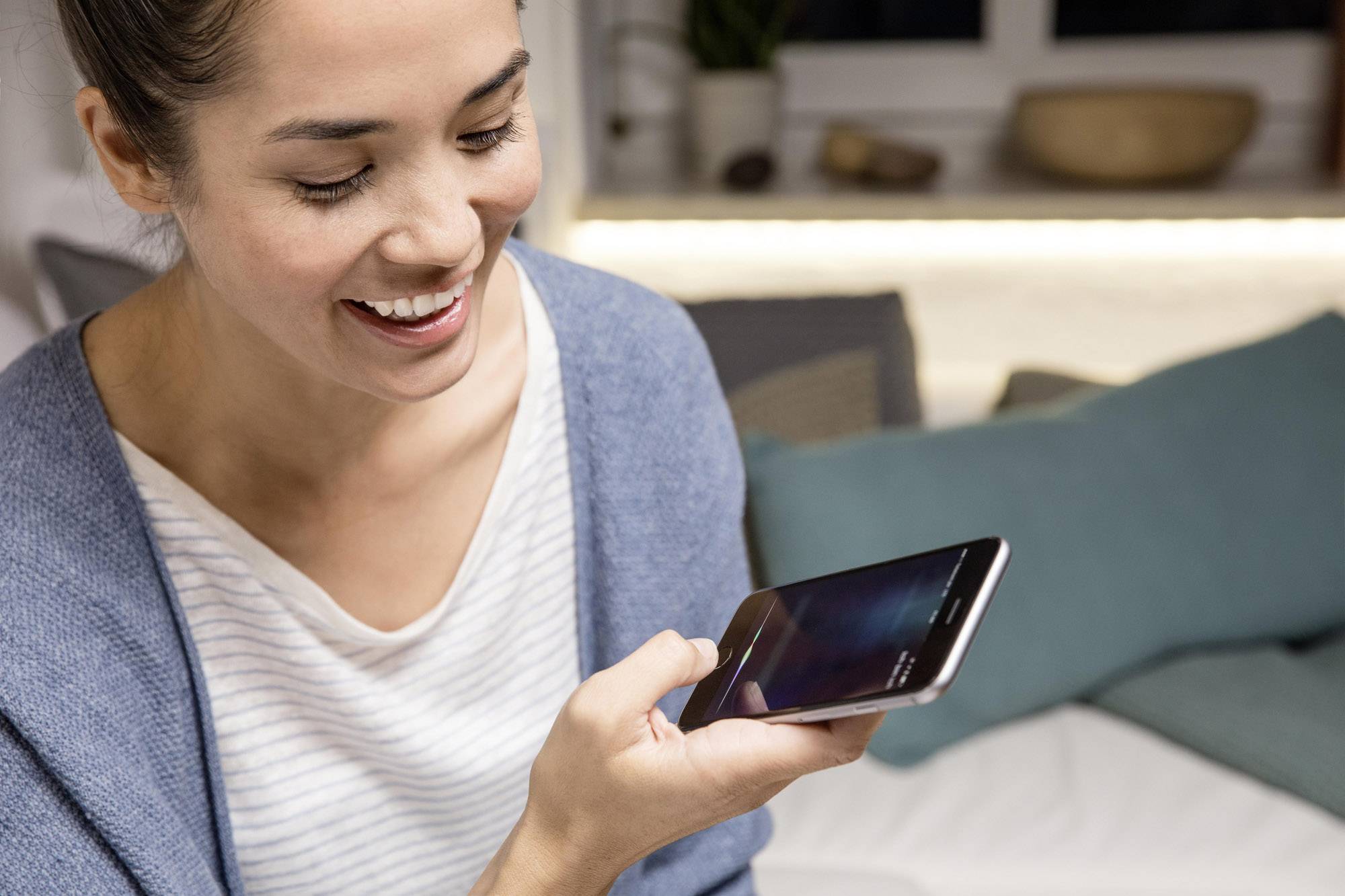 Eine lächelnde Person nutzt die Sprachsteuerungsfunktion auf einem Smartphone in einem gemütlichen Wohnzimmerumfeld.