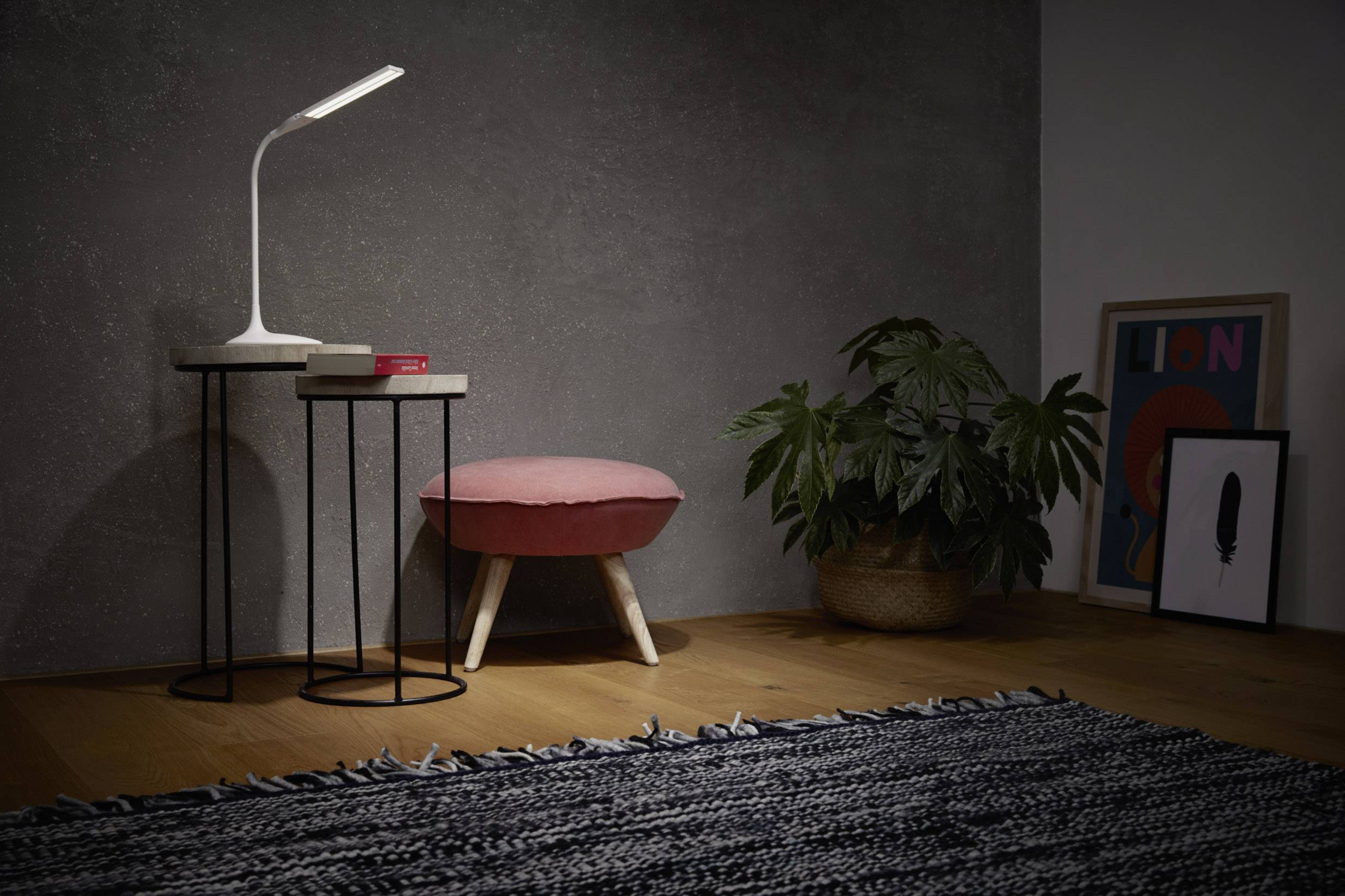Ein kleiner Tisch mit einer modernen Lampe und Büchern steht neben einem rosa Hocker. Eine Pflanze und zwei Bilderrahmen sind im Hintergrund.
