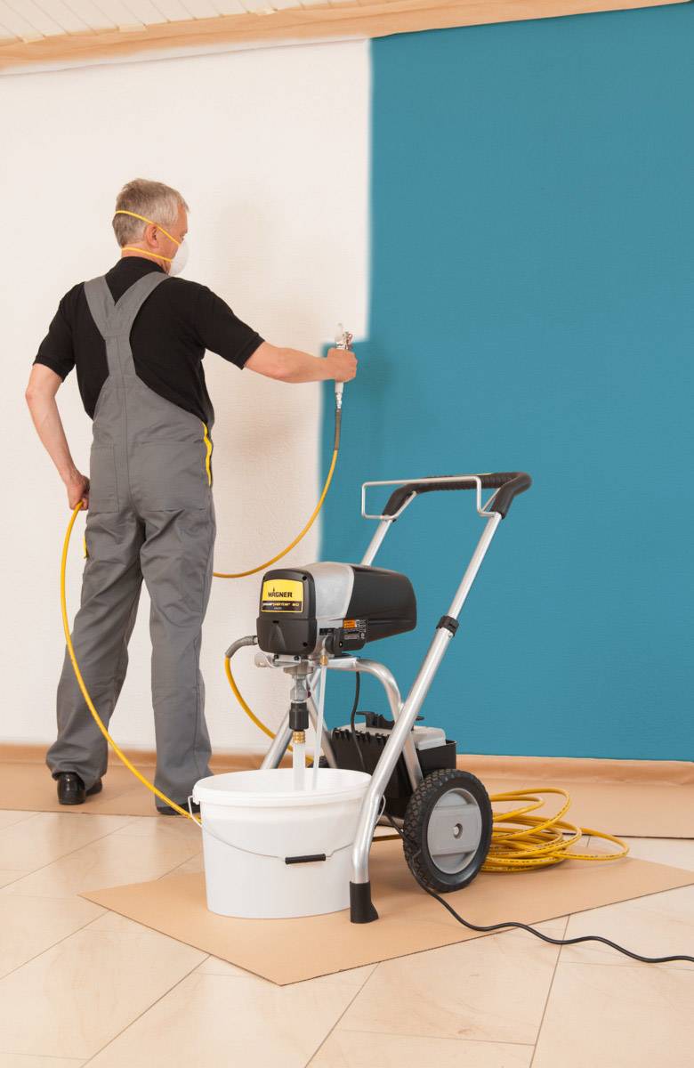 Ein Mann in Arbeitskleidung malt mit einer Farbspritzpistole eine Wand in Petrolblau an, während die andere Hälfte weiß bleibt.