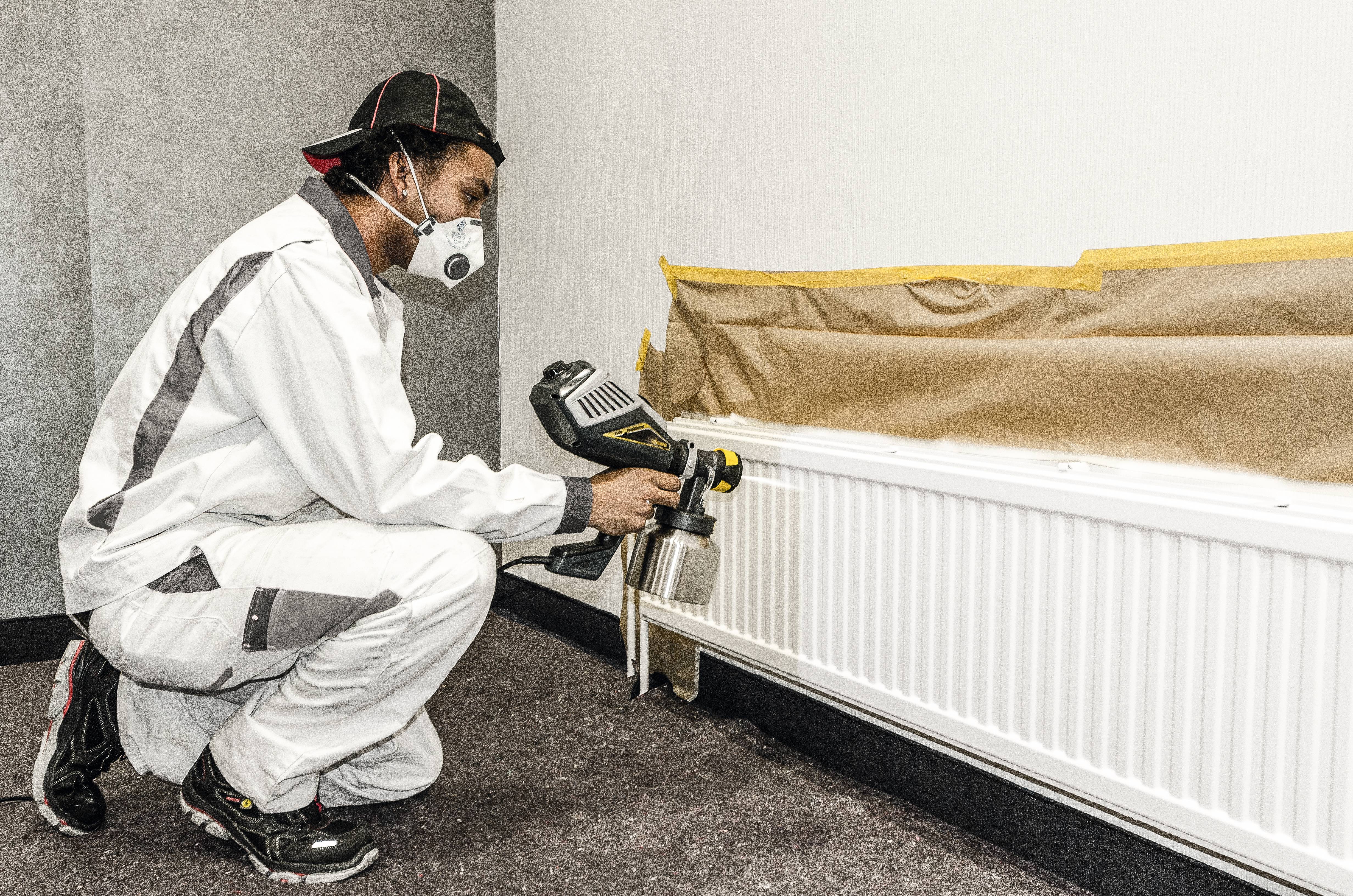 Eine Person in Schutzkleidung sprüht weiße Farbe mit einem Lackiersprüher auf einen Heizkörper. Der Bereich ist mit Papier abgedeckt.