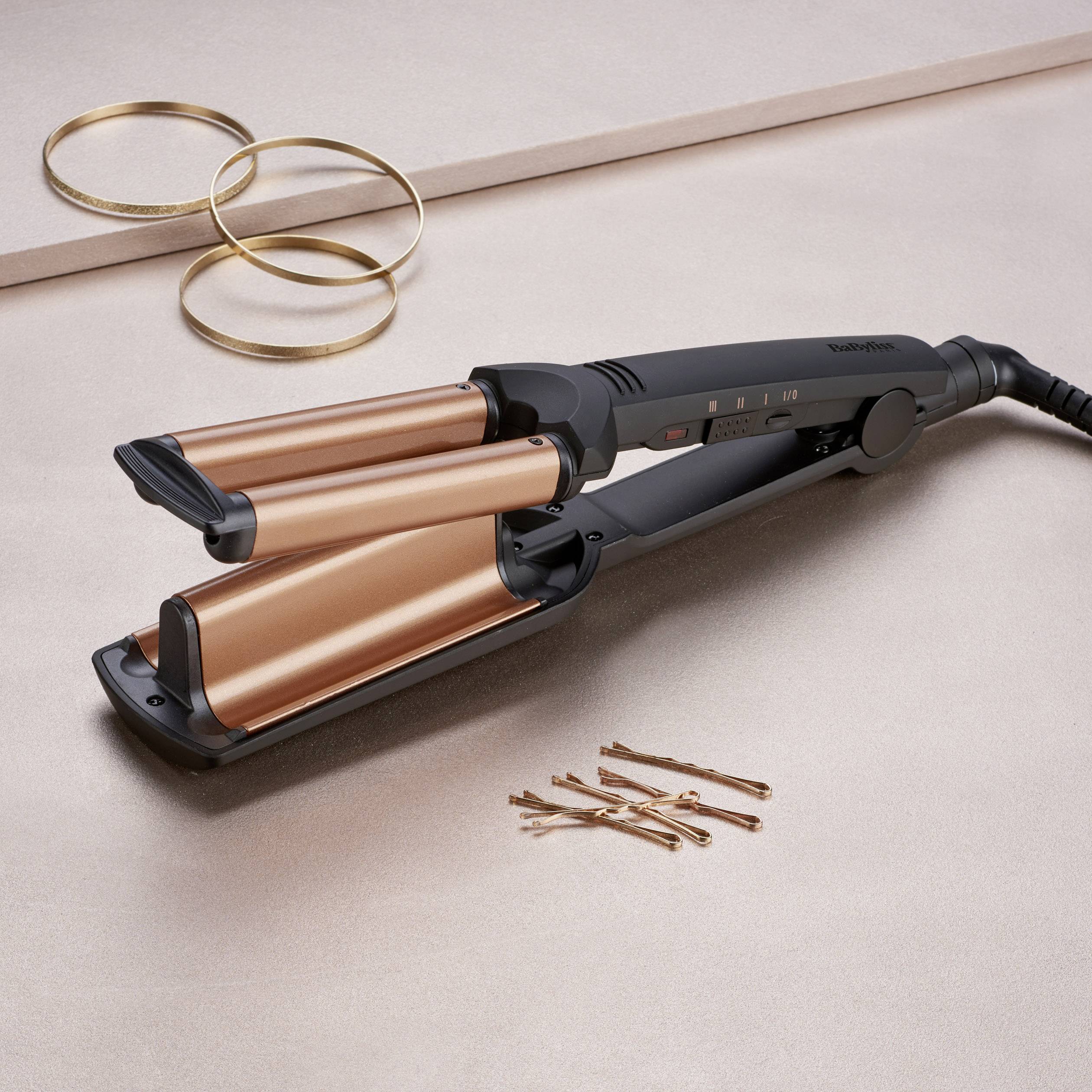Ein Lockenstab mit drei goldfarbenen Heizstäben auf einem Tisch, daneben ein paar Haarnadeln und dekorative Ringe.