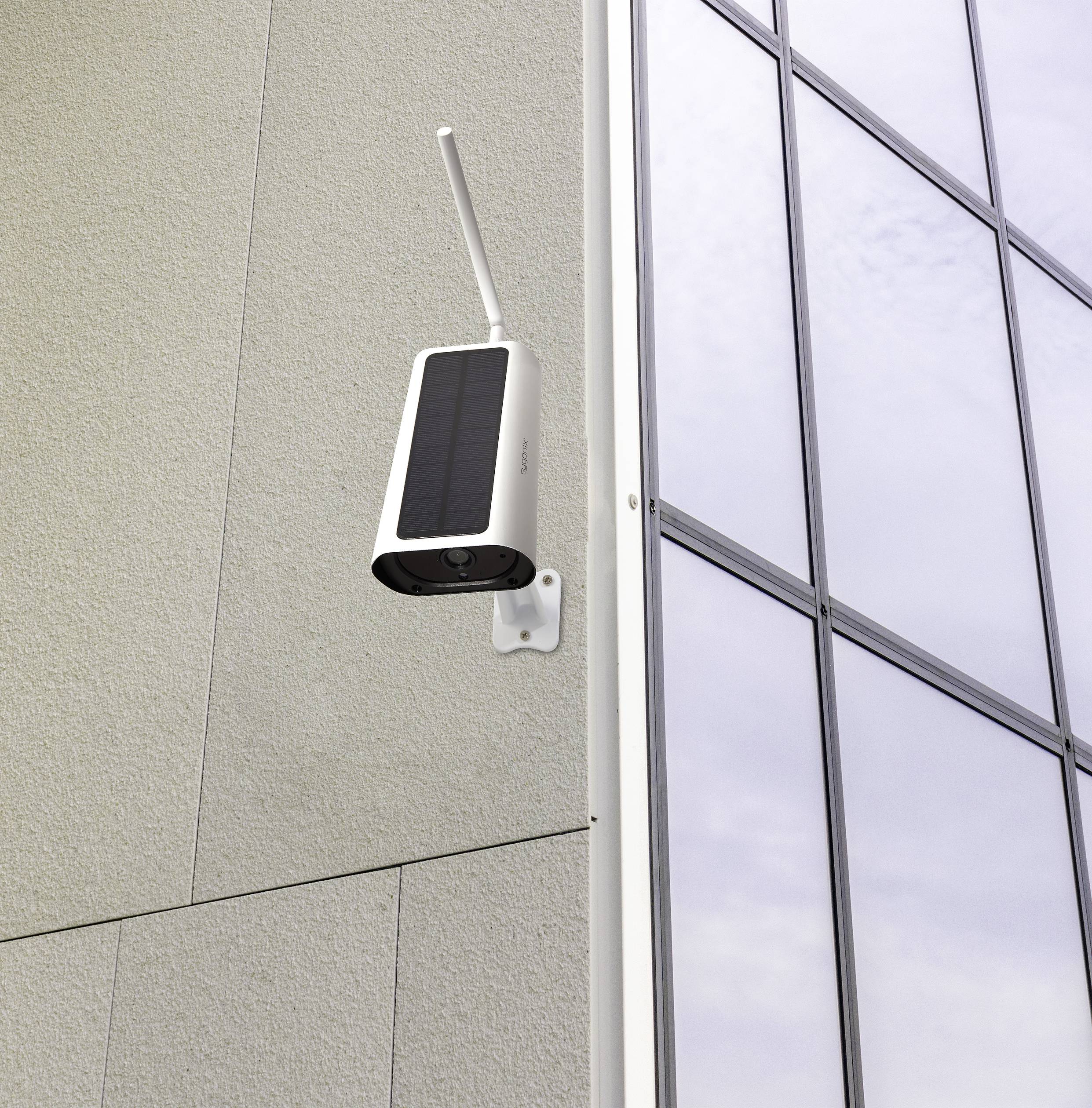 Eine Überwachungskamera mit Solarzellen montiert an einer Gebäudewand neben einem großen Fenster, zeigt Richtung Außenseite.