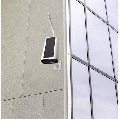 Eine Überwachungskamera mit Solarzellen montiert an einer Gebäudewand neben einem großen Fenster, zeigt Richtung Außenseite.